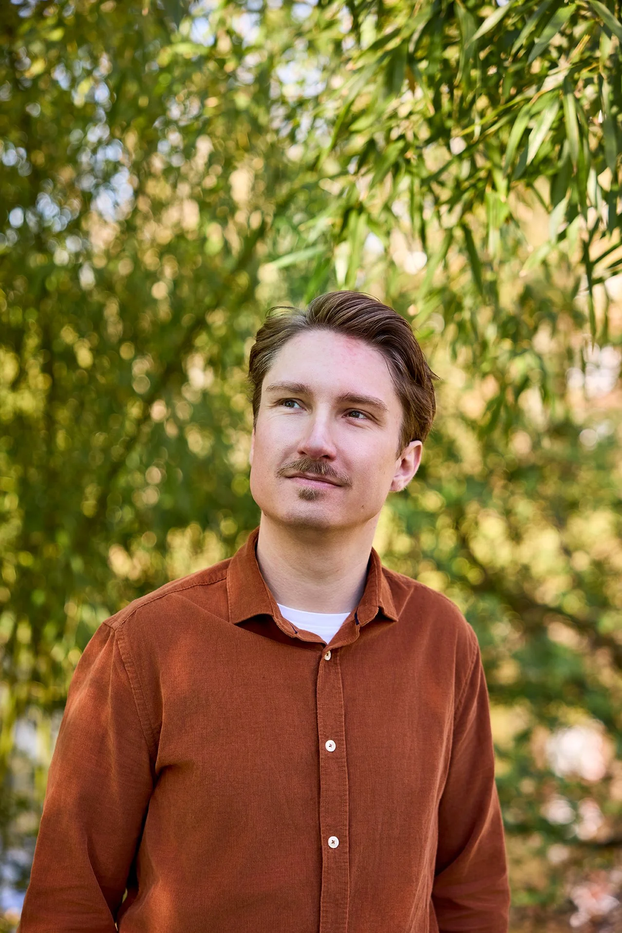 A young man with short brown hair and a mustache, wearing a brown button-up shirt, looking thoughtfully into the distance outdoors with green trees in the background.