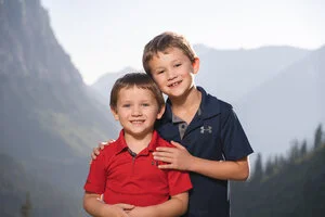 Two young boys smiling outdoors with mountains in the background.