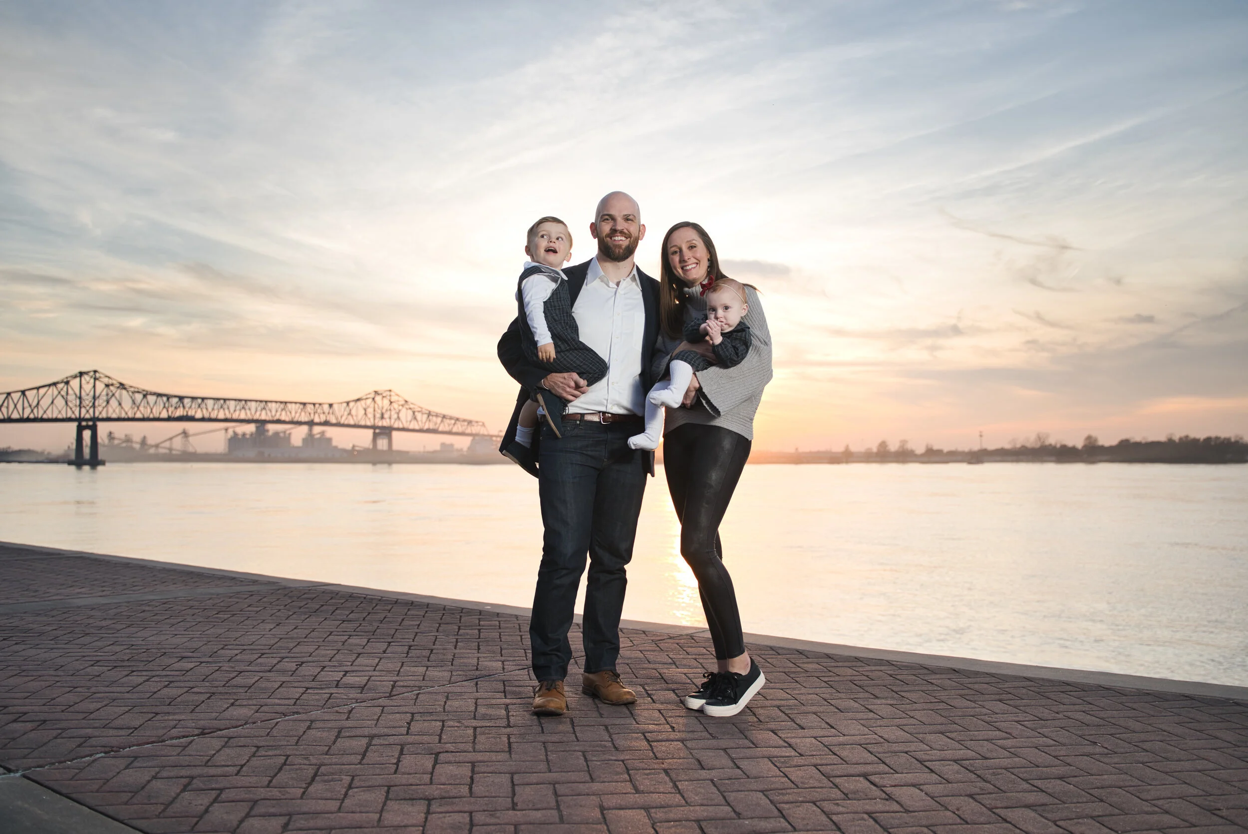 A family of four posing near a river at sunset with a suspension bridge and city skyline in the background.