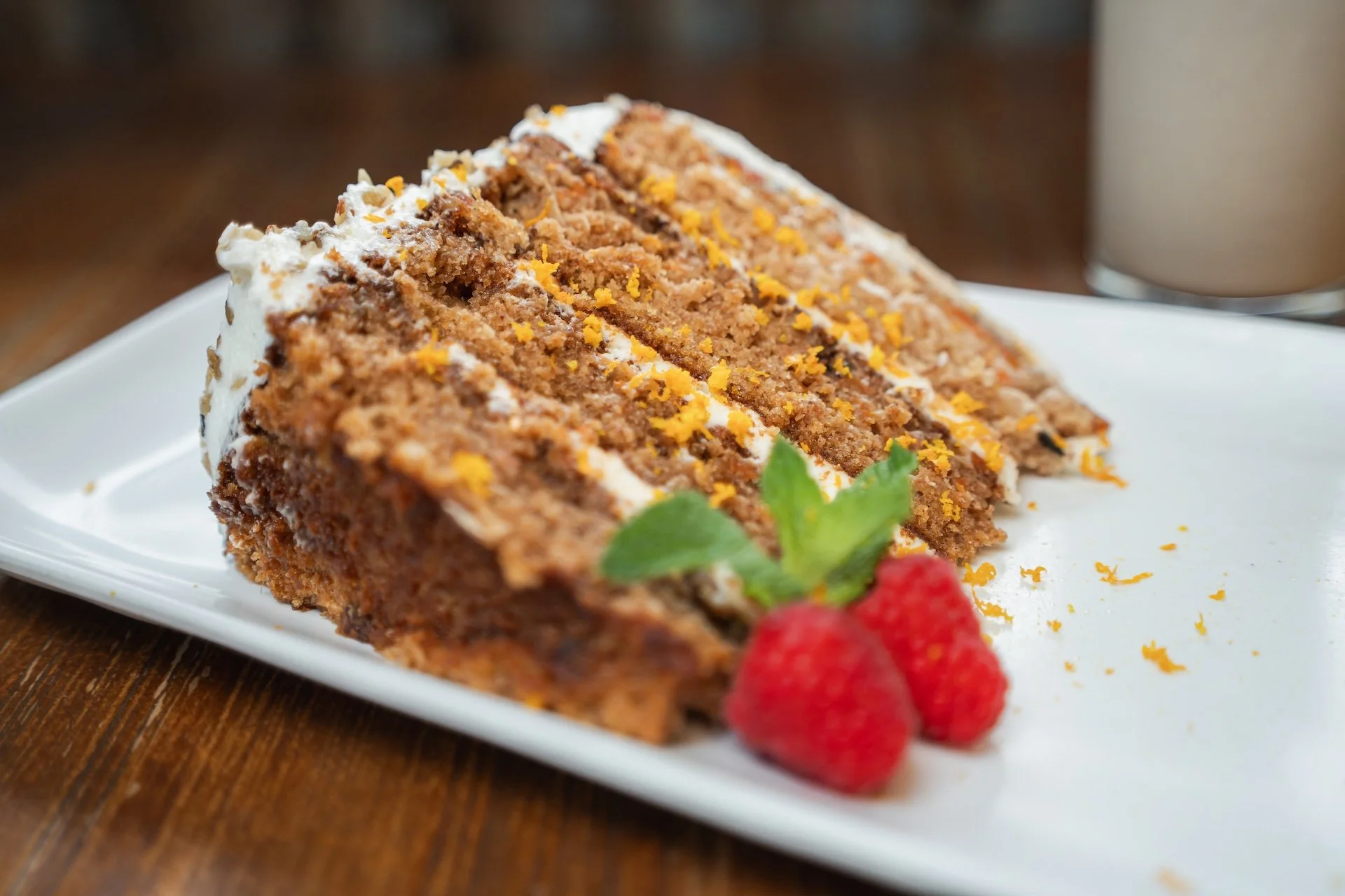 A slice of layered carrot cake with cream cheese frosting, garnished with raspberries, a mint leaf, and orange zest on a white plate.