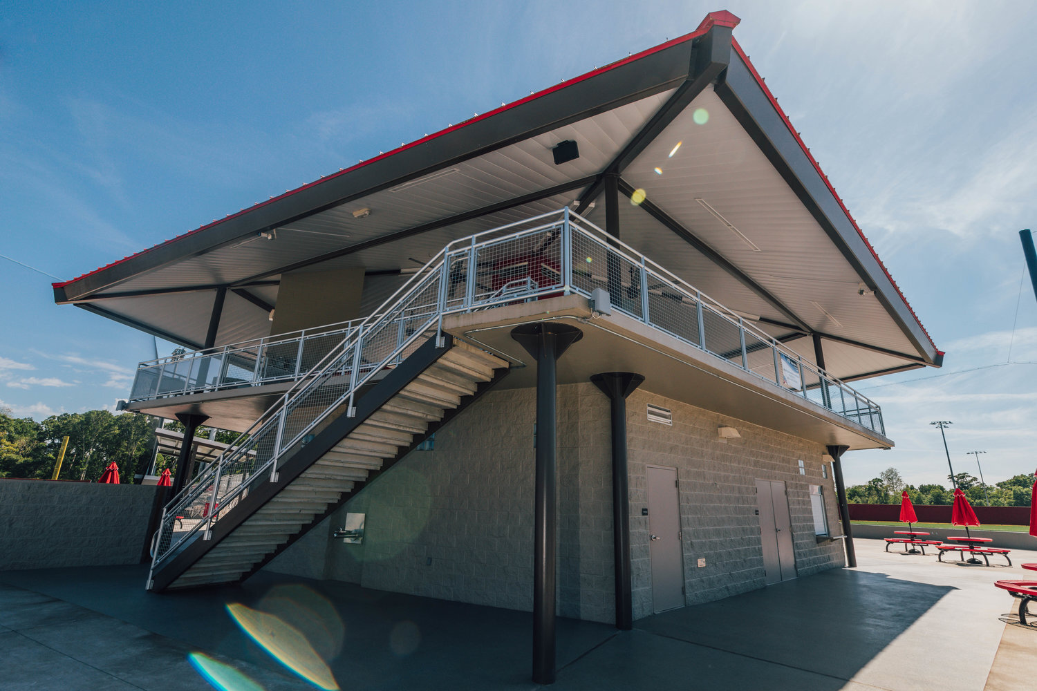 Modern building with an outdoor staircase, covered patio, and red umbrellas on picnic tables, set against a clear blue sky.