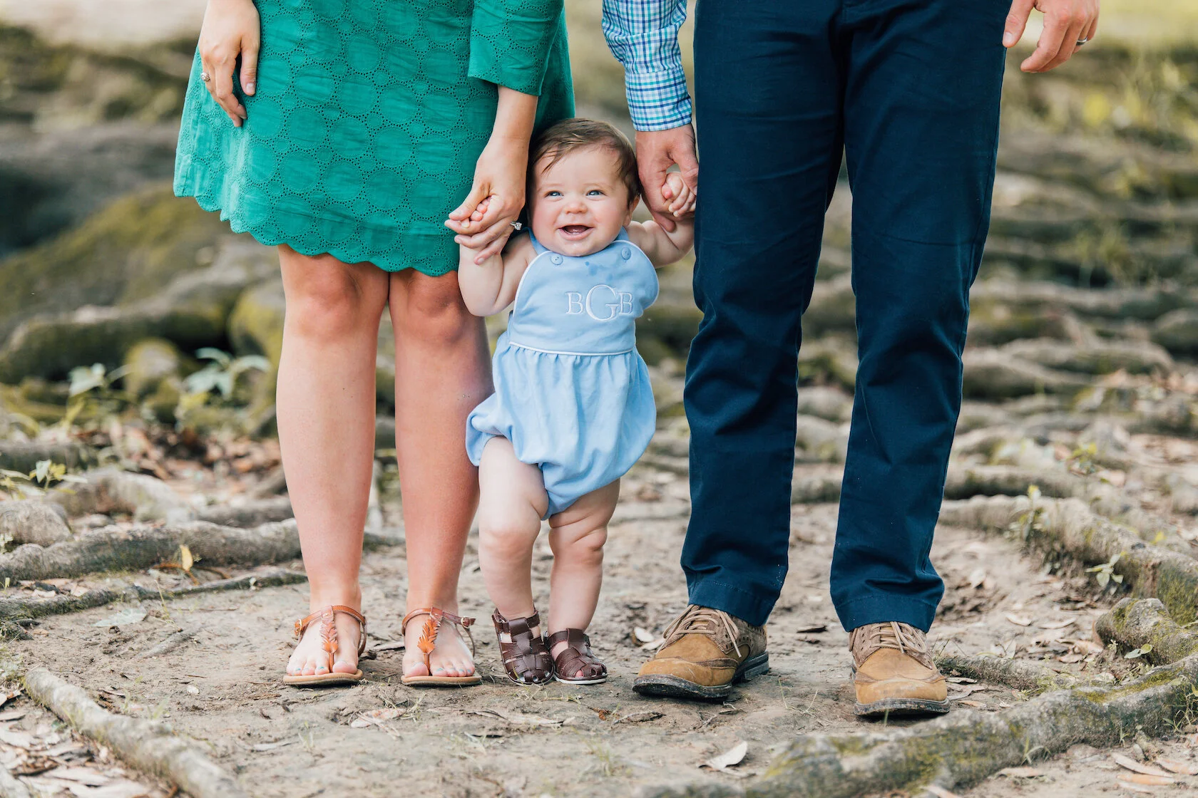 Baby in blue romper holding hands with two adults, standing on a forest trail.