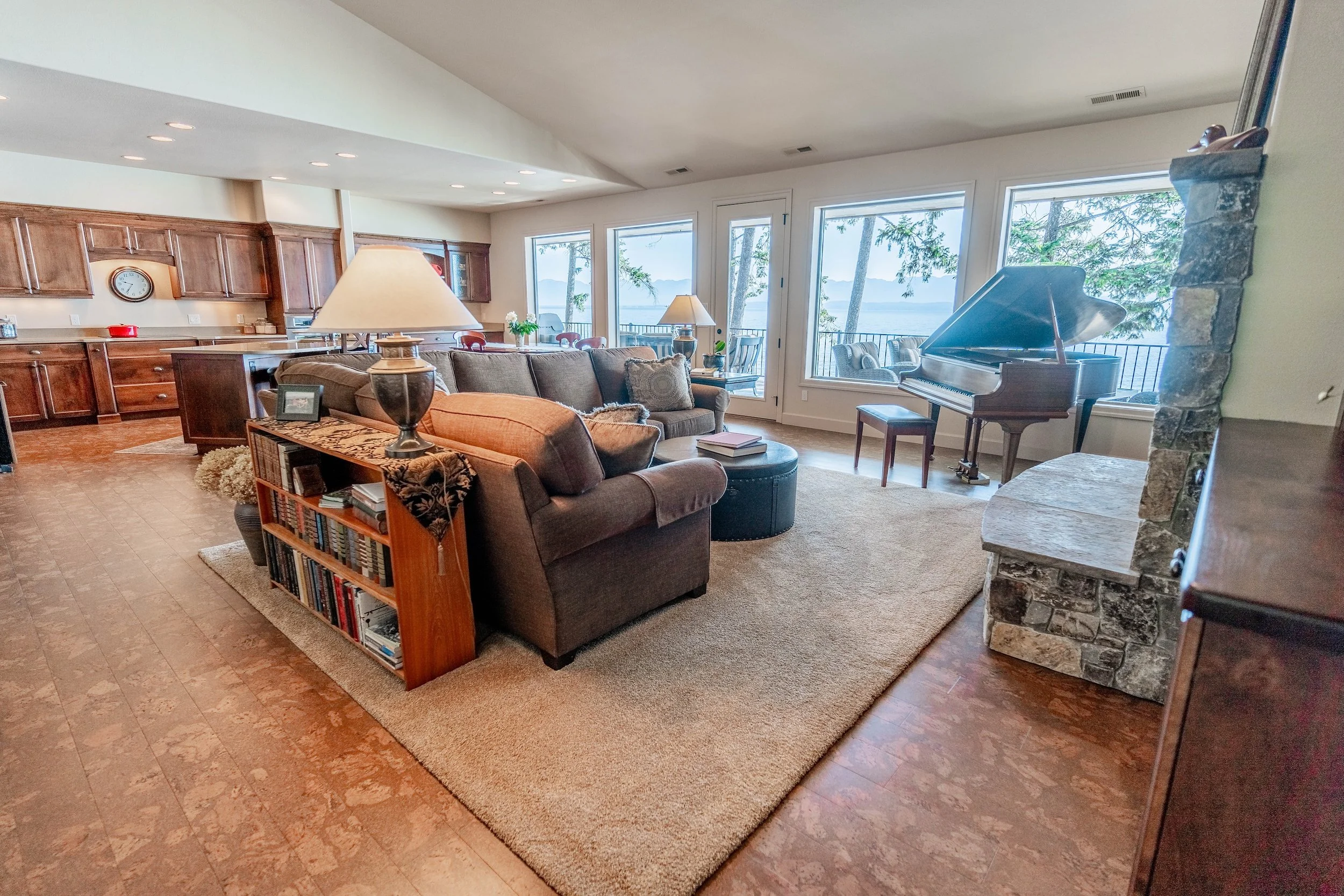 Living room with a large window view of a lake and trees, containing a traditional style grand piano, a brown sofa with cushions, a wooden bookshelf, a stone fireplace. Real Estate photographer based in Kalispell, Montana and the Flathead Valley.