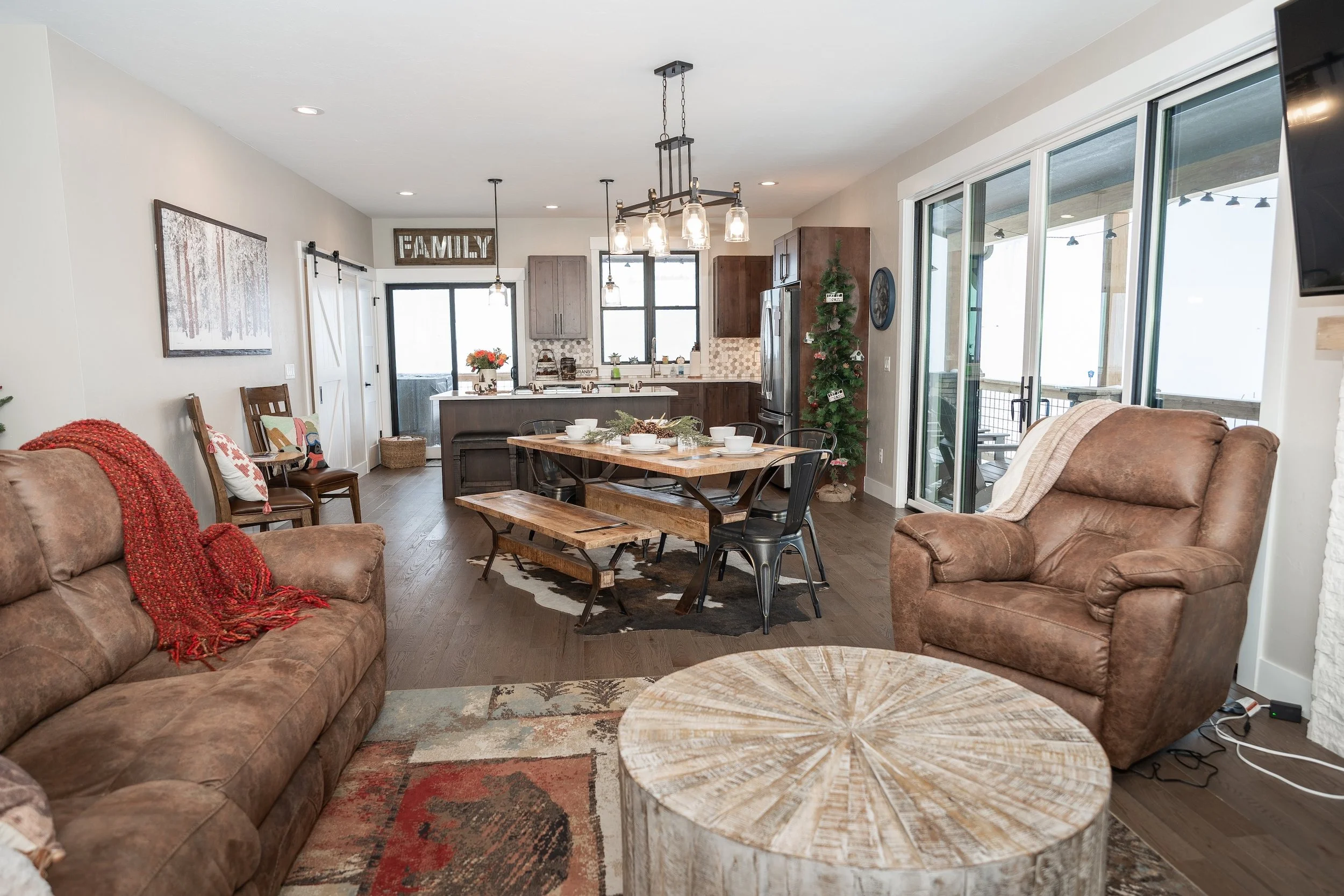 Open-concept living room and kitchen with brown leather sofas, a wooden coffee table, dining table with bowls, chairs, and a decorated Christmas tree. Real Estate photographer based in Kalispell, Montana and the Flathead Valley.