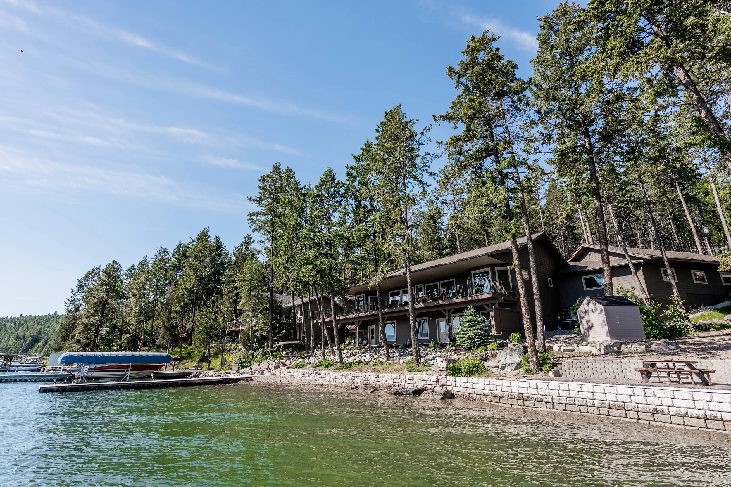 A lakeside house surrounded by tall trees, with a dock and boat in the water under a clear blue sky. Real Estate photographer based in Kalispell, Montana and the Flathead Valley.