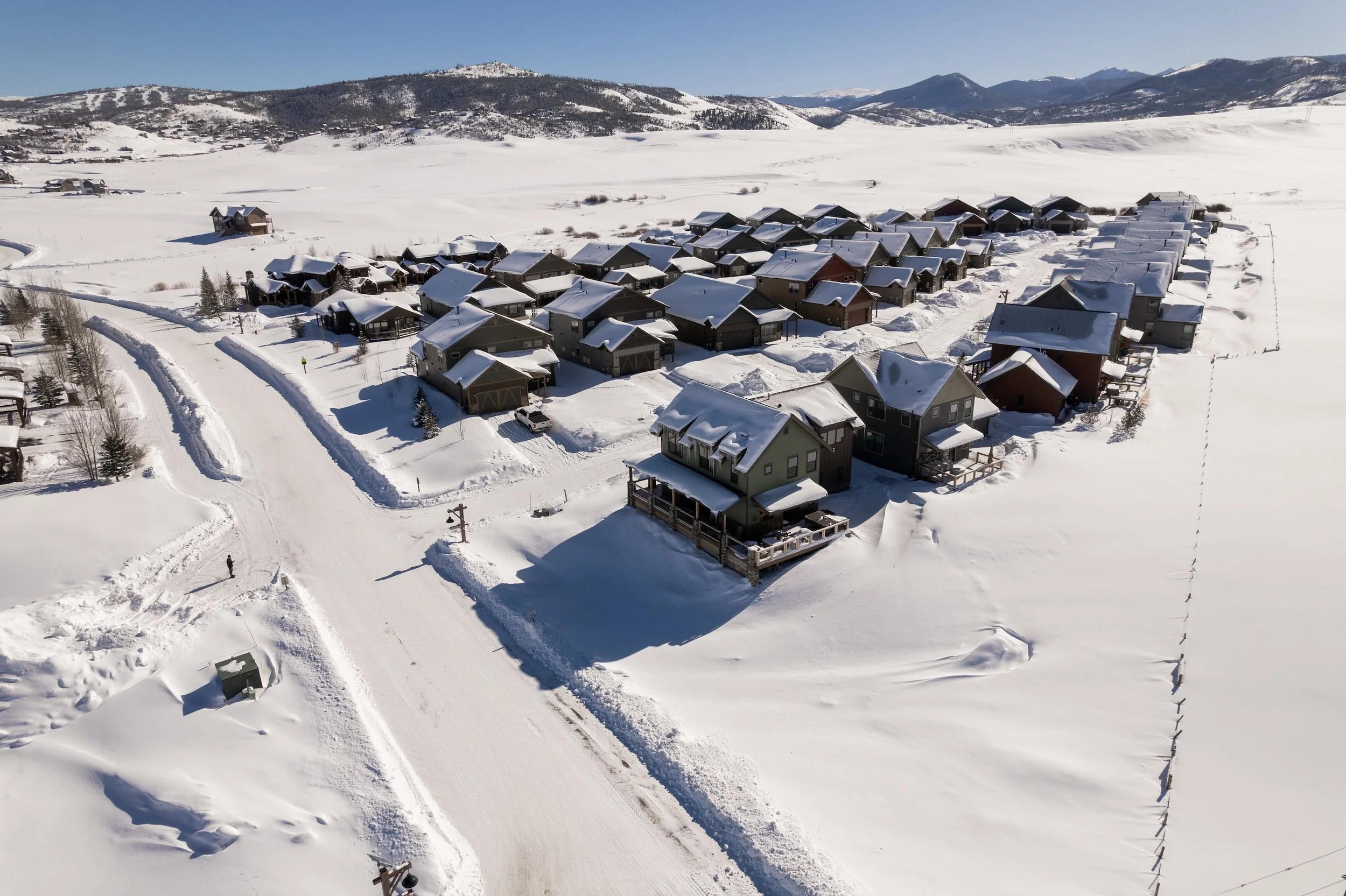 A snow-covered residential neighborhood with multiple houses, surrounded by mountains, under a clear blue sky. Real Estate photographer based in Kalispell, Montana and the Flathead Valley.