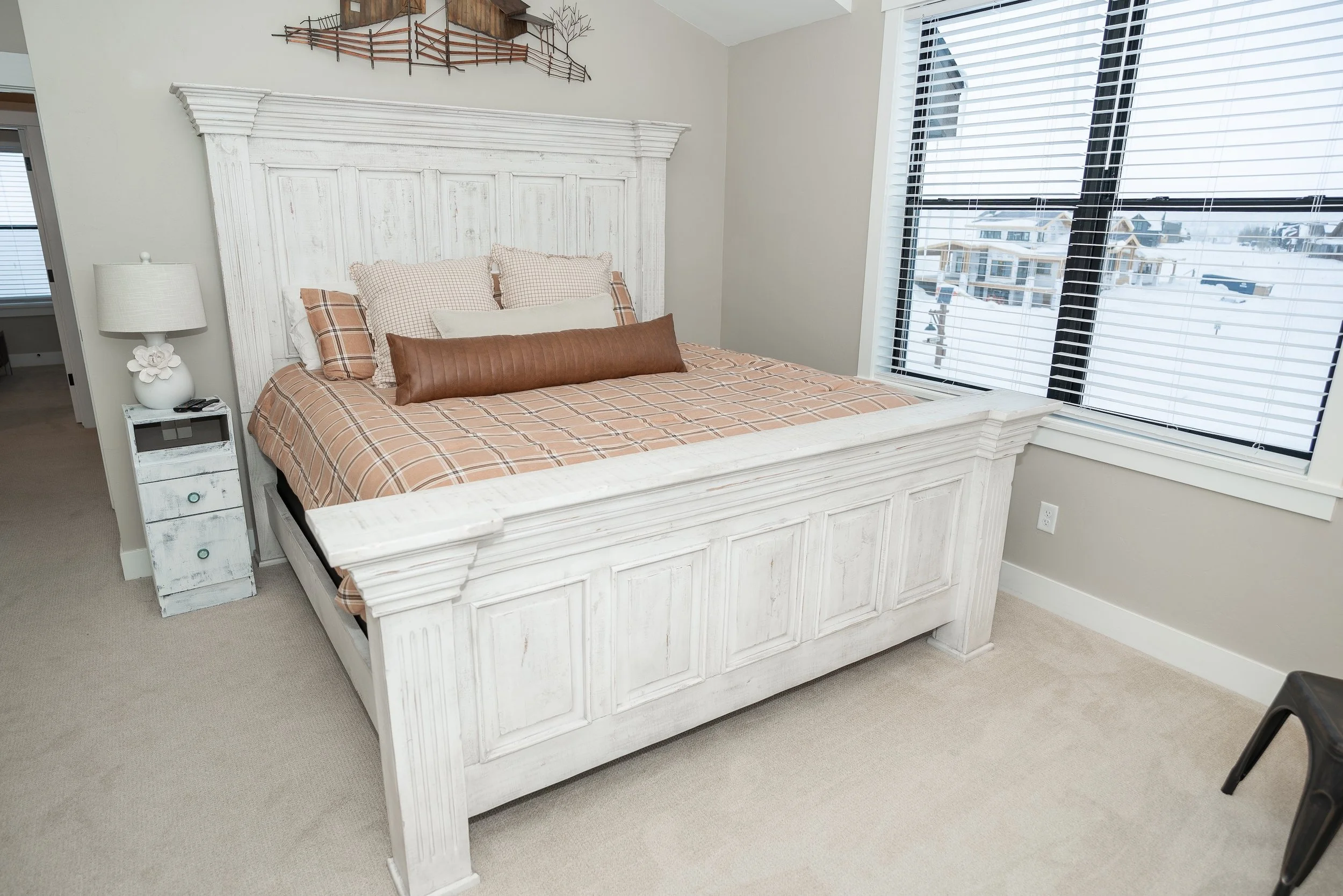 A bedroom with a white wooden bed, beige plaid bedding, multiple pillows, a white nightstand with a lamp, and a large window with blinds showing snow-covered houses outside. Real Estate photographer based in Kalispell, Montana and the Flathead Valley