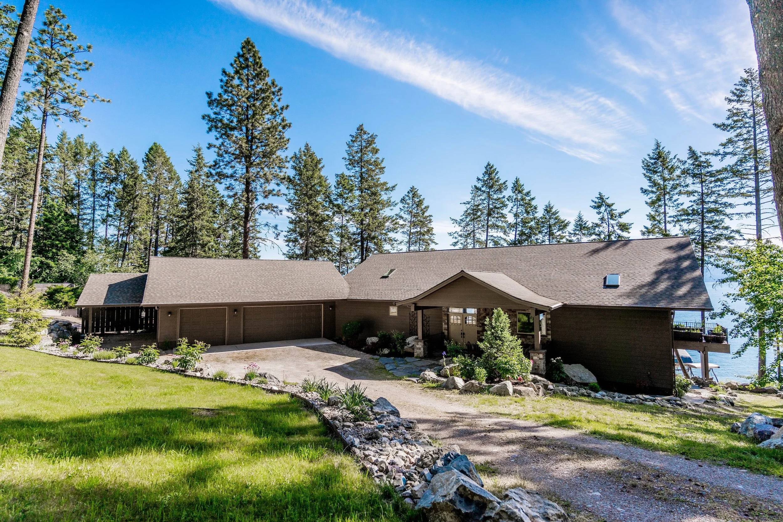 A house with a sloped roof surrounded by tall trees, green grass, and rocks on a bright, sunny day. Real Estate photographer based in Kalispell, Montana and the Flathead Valley.