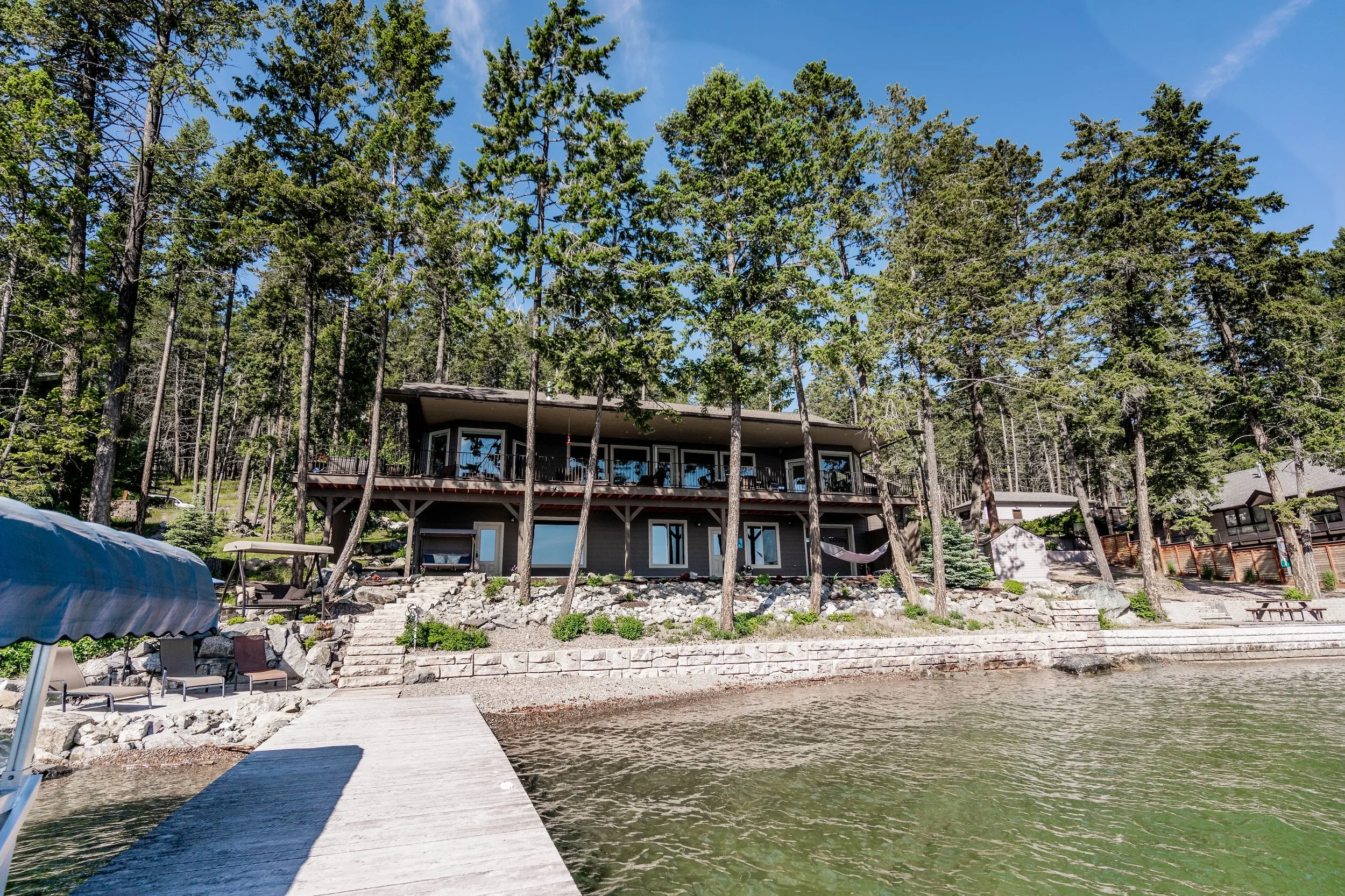 A lakeside house on a hill surrounded by trees, with a dock extending into the water, and outdoor furniture laid out on the shore. Real Estate photographer based in Kalispell, Montana and the Flathead Valley.