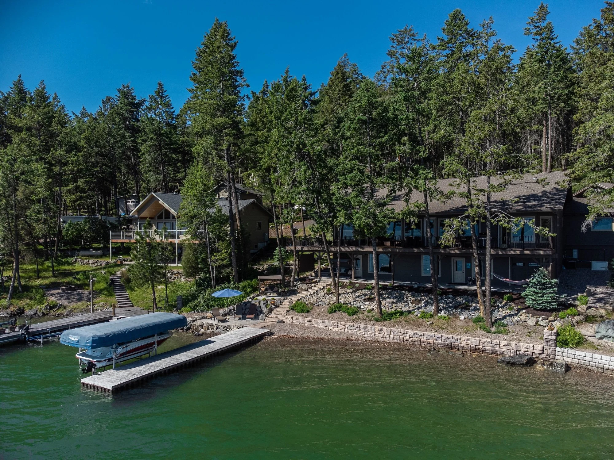 A lakeside view showing a house with a large deck among tall pine trees, a dock with a boat covered by a sunshade, Real Estate photographer based in Kalispell, Montana and the Flathead Valley.