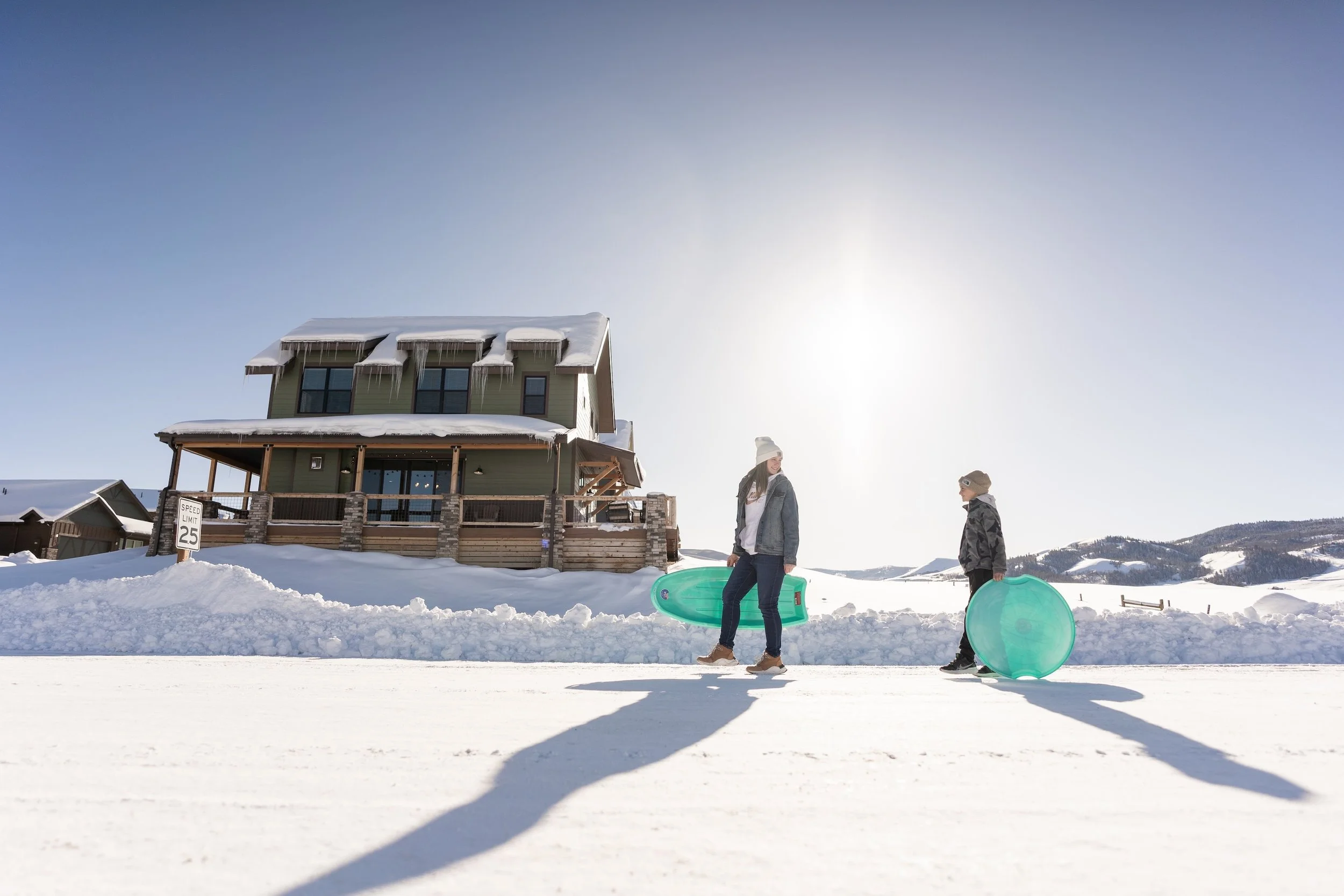 A woman and a young boy walking on snow-covered ground, each carrying a transparent sled, with a large snow-covered house and mountains in the background under bright sunlight.