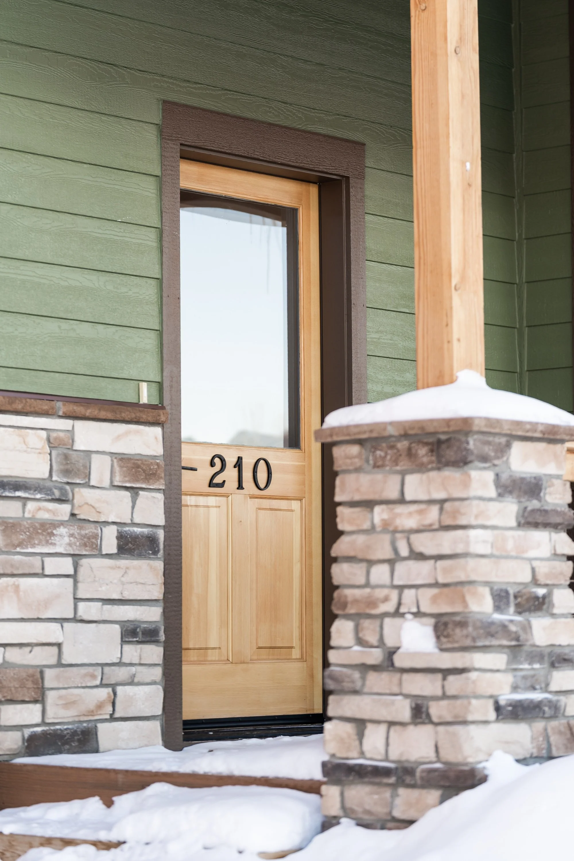 Close-up of a wooden front door with house number 210, flanked by stone columns, with snow on the ground. Real Estate photographer based in Kalispell, Montana and the Flathead Valley.