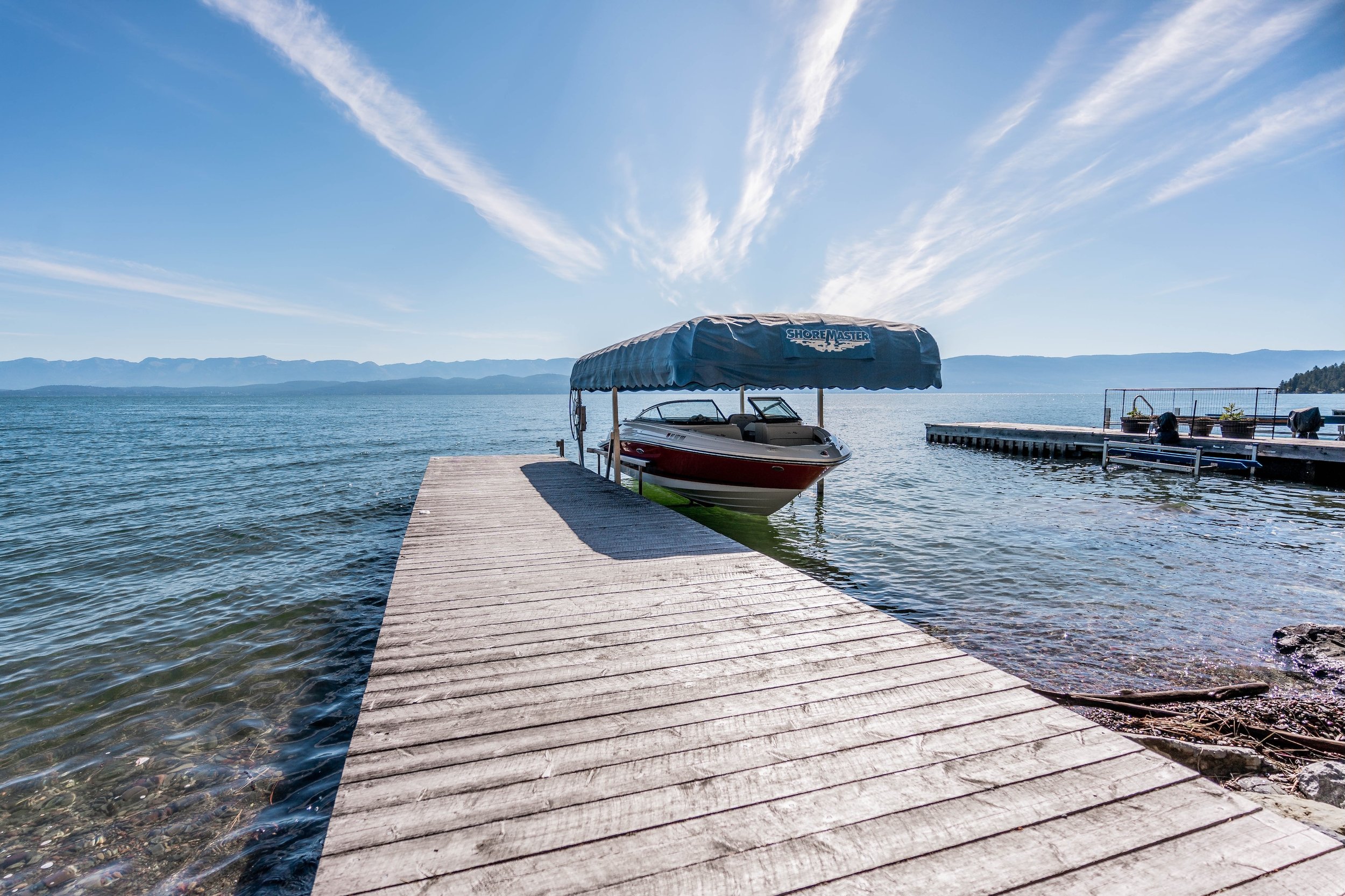 A wooden dock extends into a lake with a boat under a blue canopy, and a second dock with plants and equipment, with mountains in the background and a partly cloudy sky. Real Estate photographer based in Kalispell, Montana and the Flathead Valley.