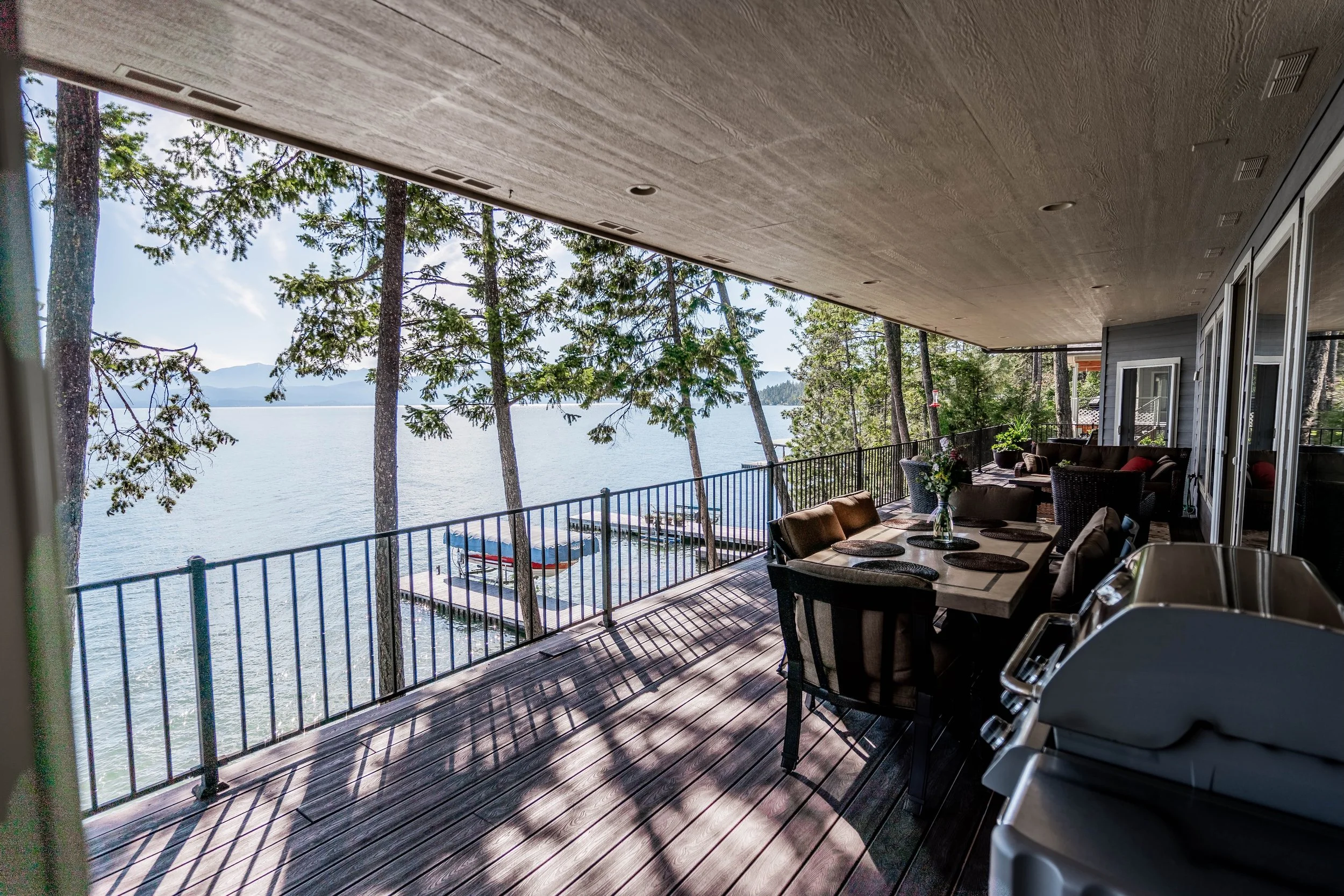 Covered balcony overlooking a lake with trees and boats on a dock, furnished with outdoor chairs, Real Estate photographer based in Kalispell, Montana and the Flathead Valley.
