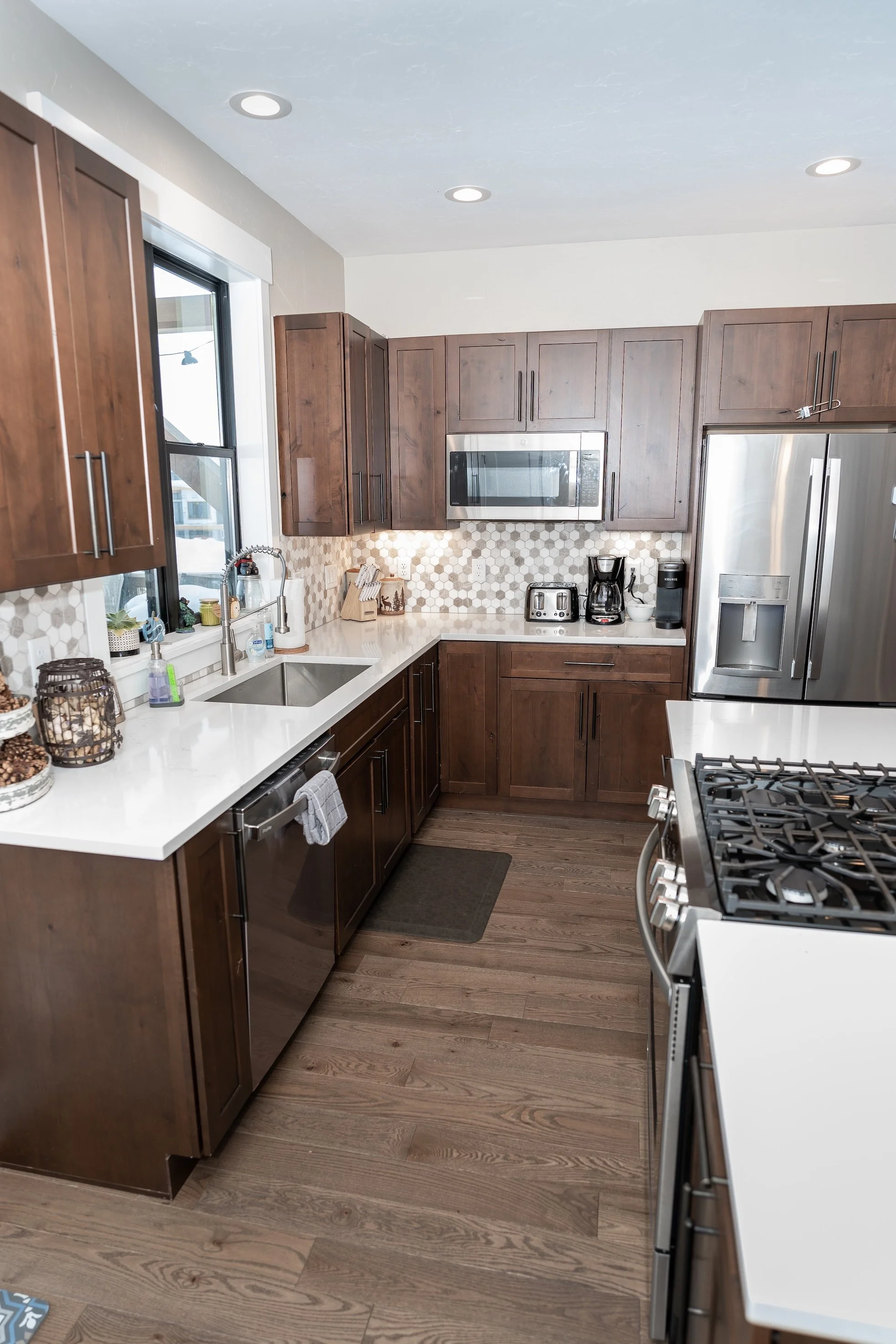 A modern kitchen with dark wood cabinets, white countertops, stainless steel appliances including a refrigerator, microwave, and stove, and a small window above the sink. Real Estate photographer based in Kalispell, Montana and the Flathead Valley.