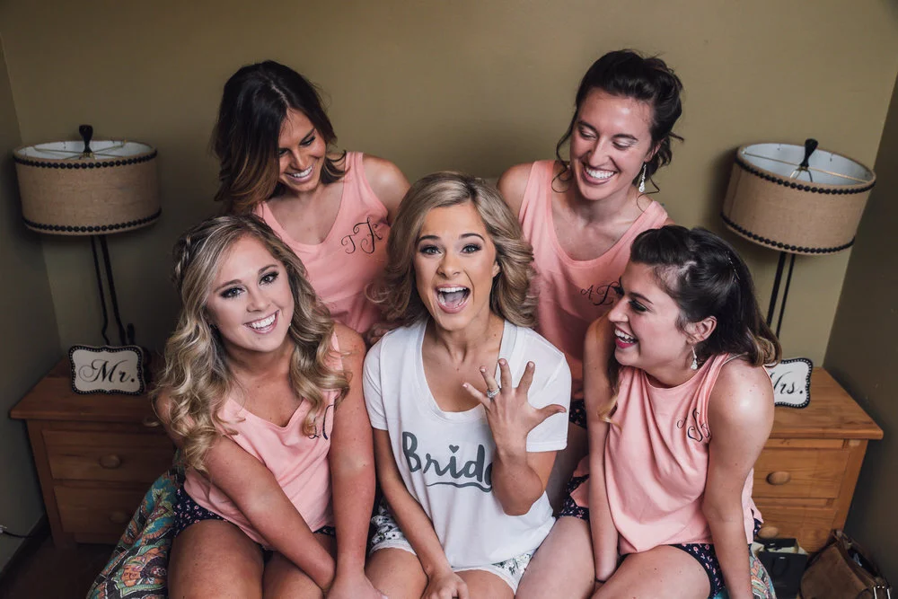 A group of five women sitting on a bed, with one woman in the center wearing a "Bride" shirt and displaying an engagement ring. The others wear matching pink shirts and are smiling and laughing. The setting includes two lamps and signs reading "Mr."