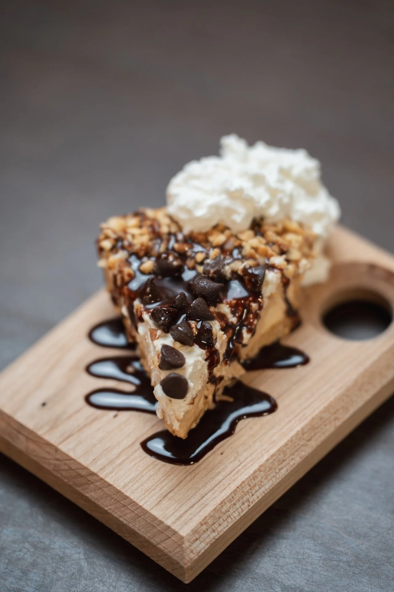 A slice of cheesecake topped with whipped cream, chocolate chips, and chopped nuts on a wooden serving board with chocolate syrup drizzled around it.