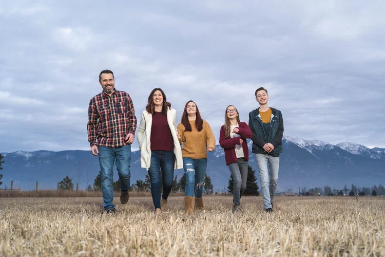 A family of five walking in a field with mountains in the background. The group includes three adults and two children, all wearing casual winter clothing. The sky is overcast, and the landscape is open and natural.