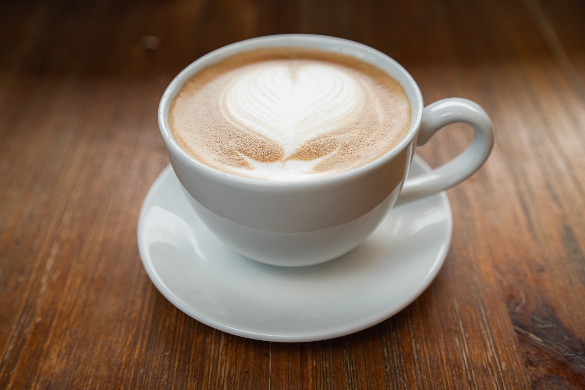 A white ceramic cup filled with latte featuring a heart-shaped latte art on top, placed on a matching saucer on a wooden table.