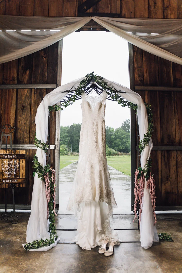 Wedding dress hanging in barn doorway, adorned with greenery and draped fabric, with outdoor scenery visible through the entrance.