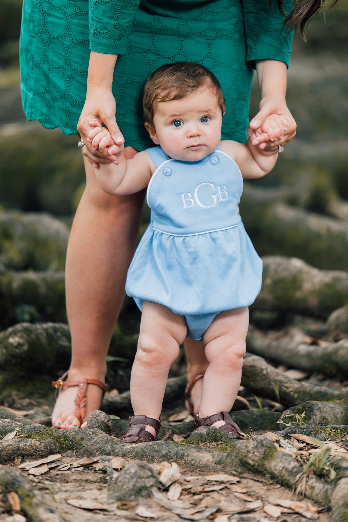 A baby wearing a blue outfit with initials 'BGB' being held by an adult in a green dress, outdoors on tree roots.