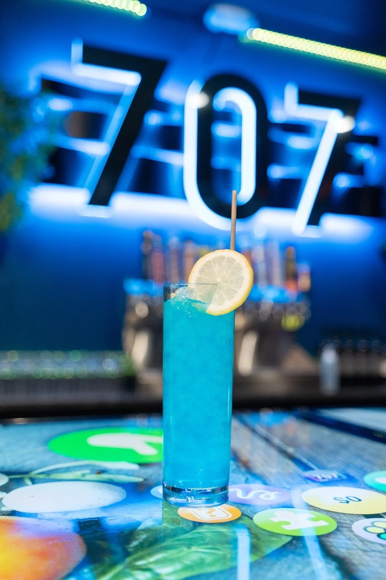 A tall glass of blue cocktail garnished with a lemon slice and a stir stick, set on a colorful table in front of a blue neon sign that says 'Zoz' at a bar.