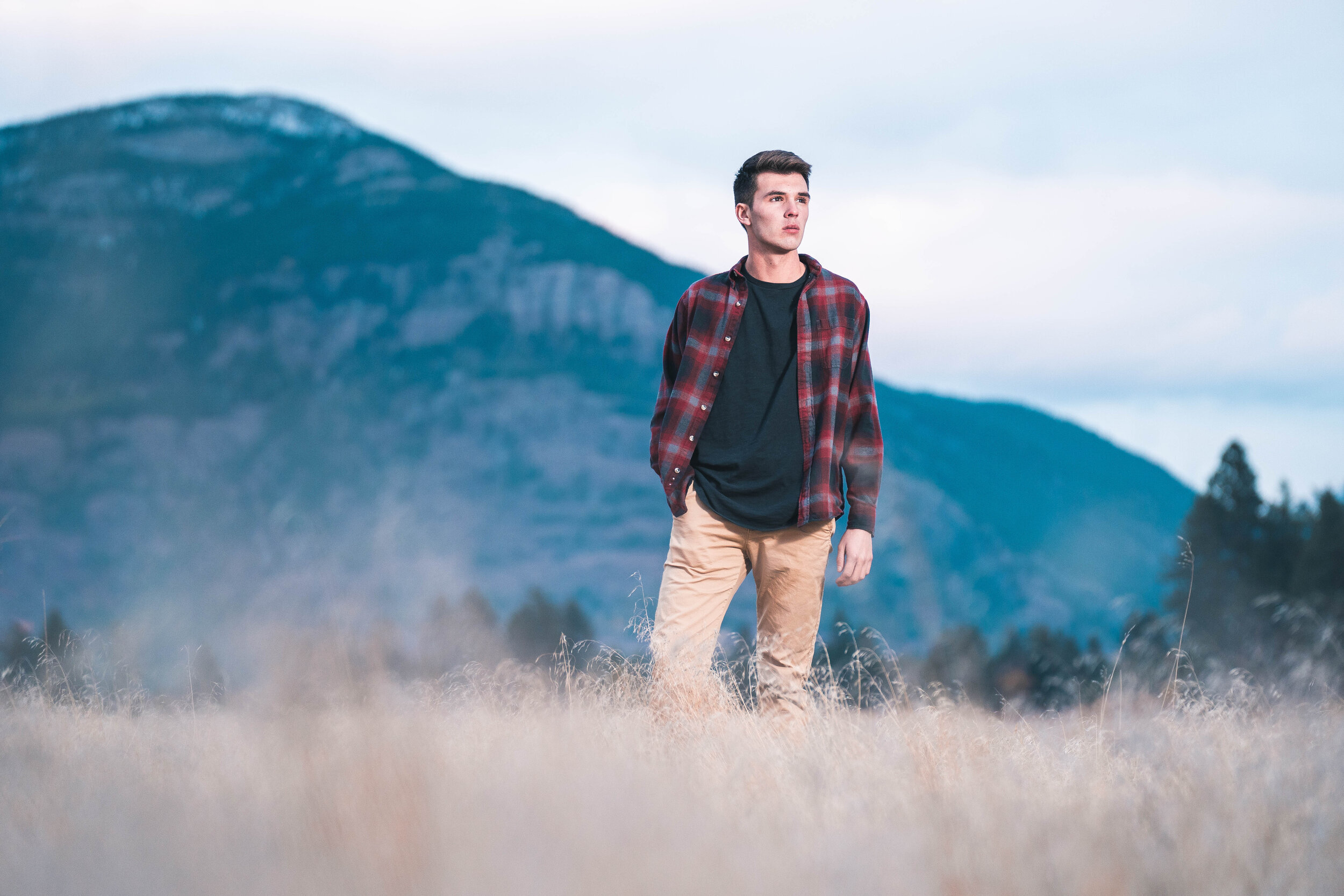 Man in plaid shirt walking in grass field with mountains in the background.