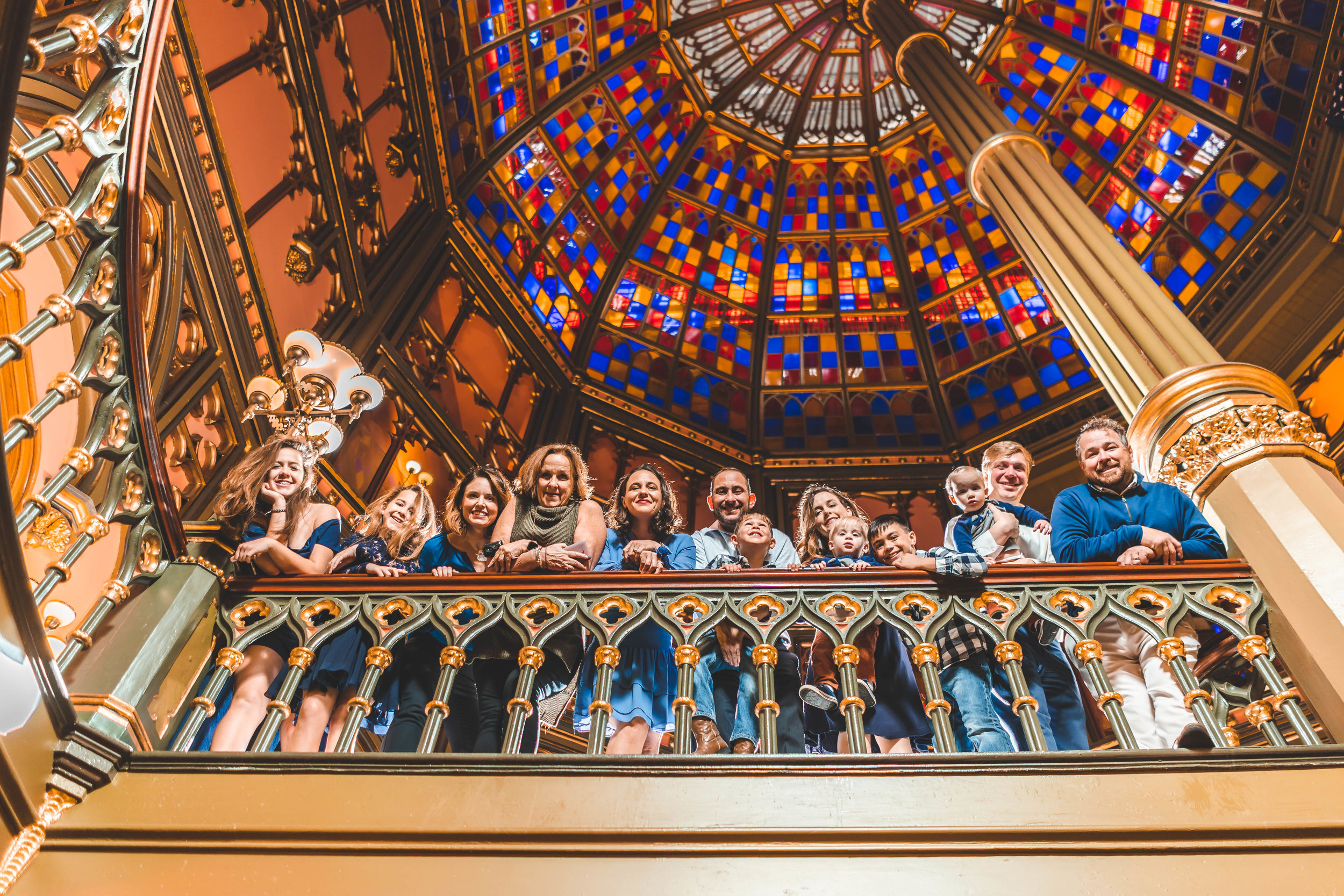 A diverse group of people standing on a decorative balcony under a colorful stained glass ceiling. They appear to be smiling and enjoying the architectural setting.