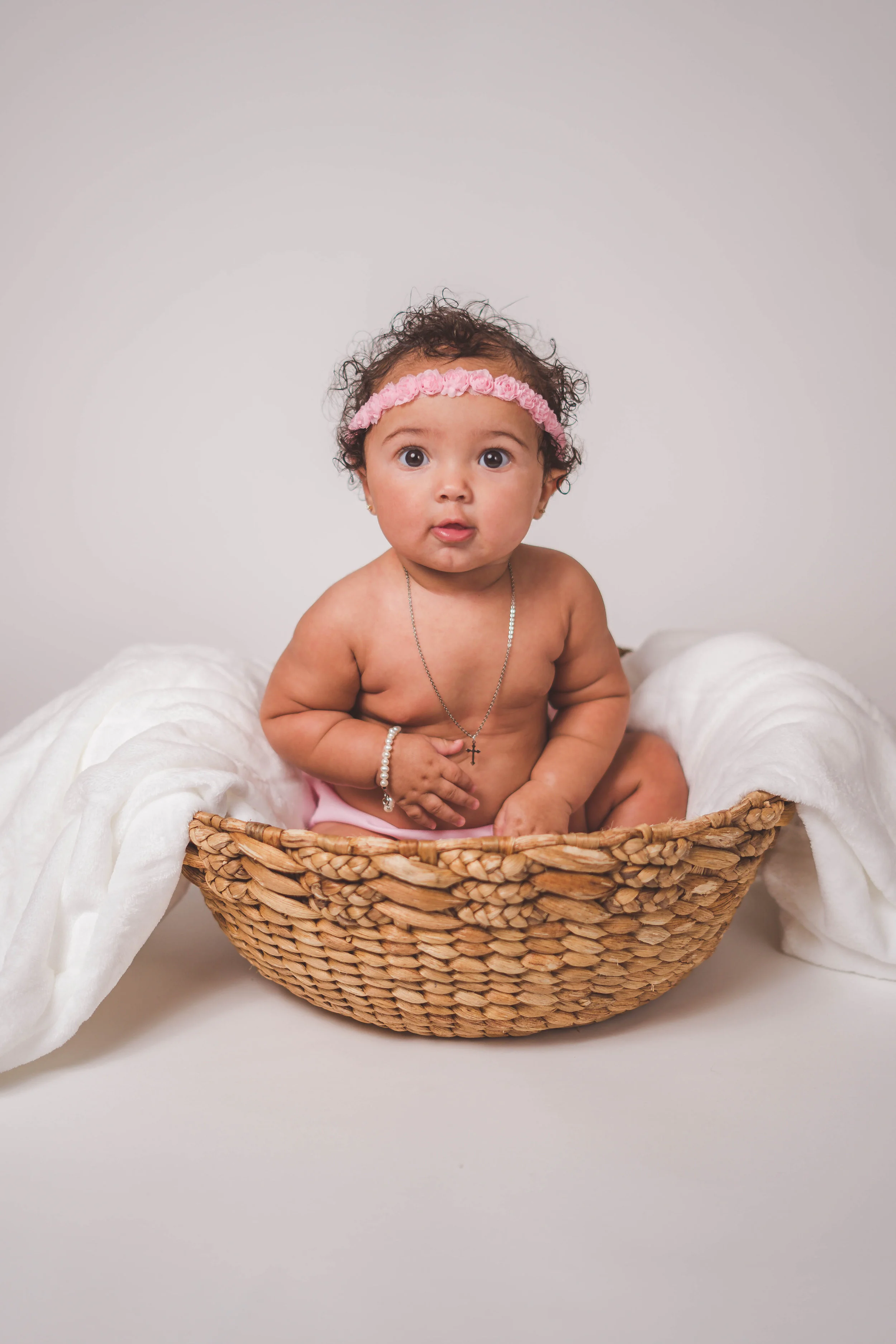 A baby sitting in a woven basket, wearing a pink headband and a necklace with a cross pendant, on a light background with a white blanket.