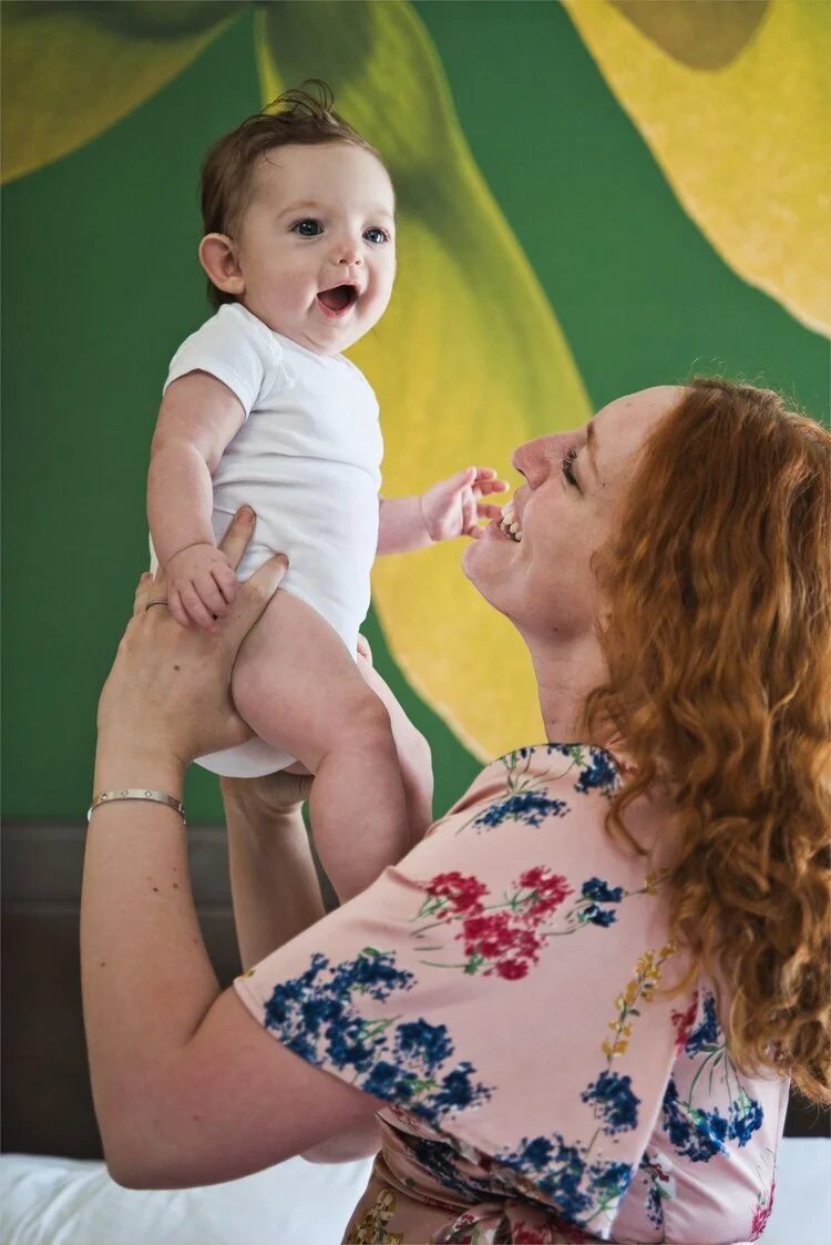 Woman holding a smiling baby in a white onesie, in front of a green and yellow background.