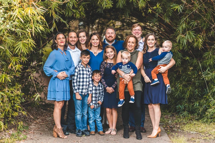 A large family group posing outdoors in front of a leafy arch, with some members wearing blue attire. There are adults, children, and toddlers smiling together.