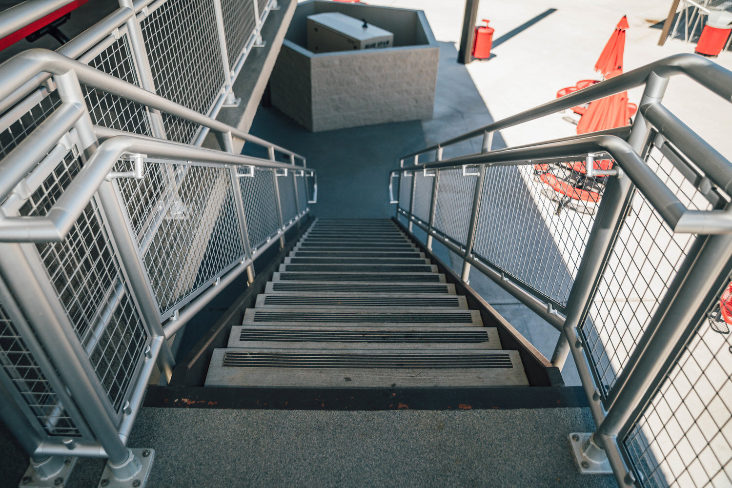 Metal staircase with railings and mesh panels, leading downward to an outdoor area with red umbrellas and tables in the distance.