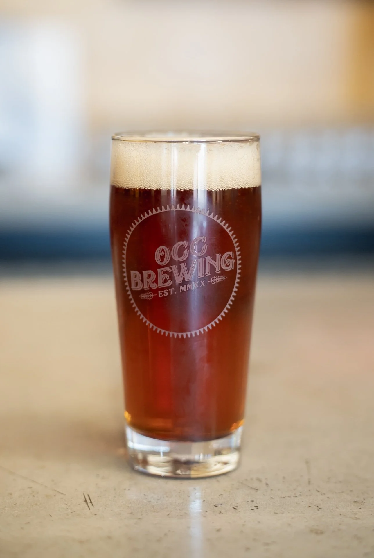 A pint glass with OC Brewing logo, filled with amber-colored beer with a foamy head, on a light-colored surface.