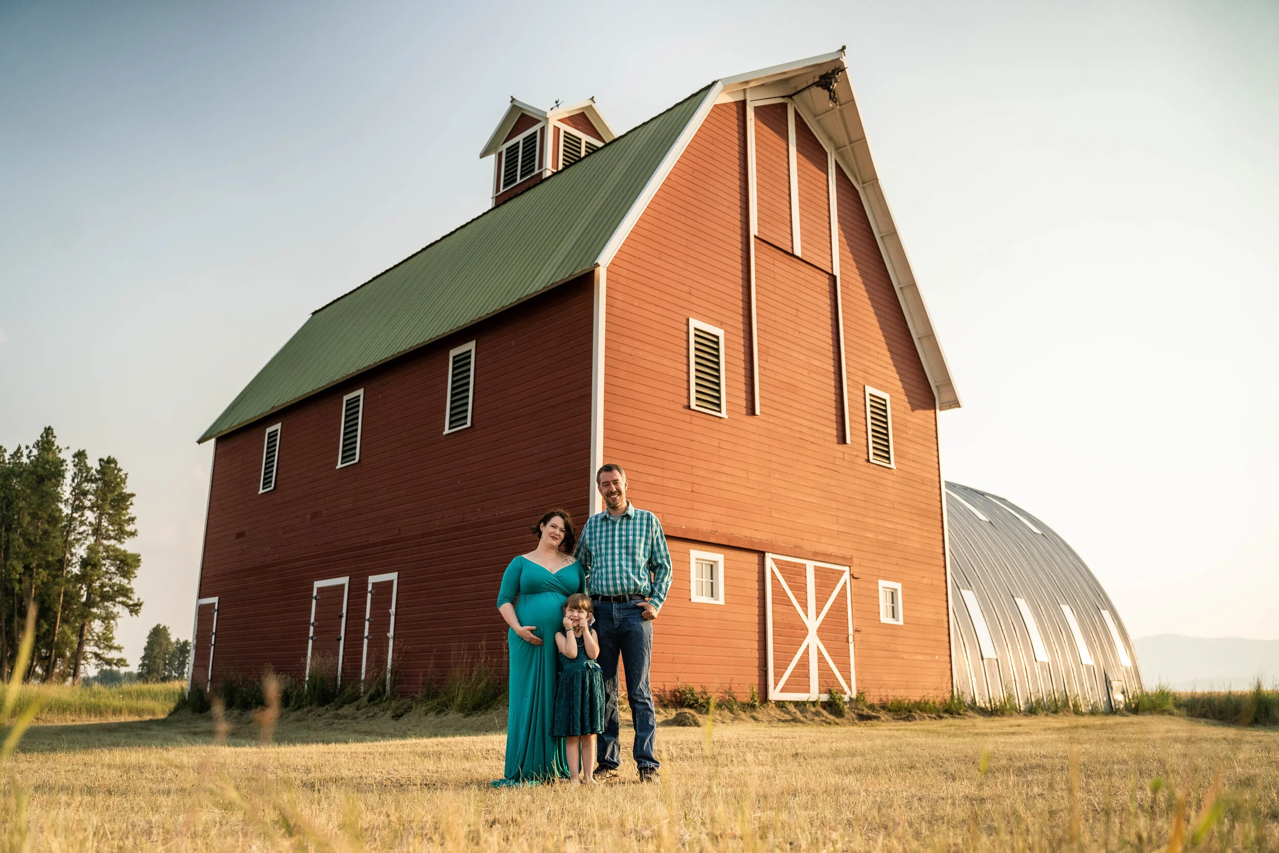 A family standing in front of a large red barn with a green roof in a rural setting. The family includes a pregnant woman in a teal dress, a man in a plaid shirt, and a young girl in a dress.