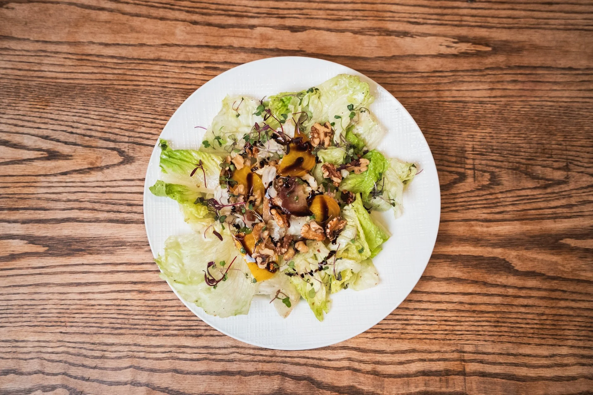 Salad with lettuce, microgreens, walnuts, chopped vegetables, and balsamic glaze on a white plate on a wooden table.