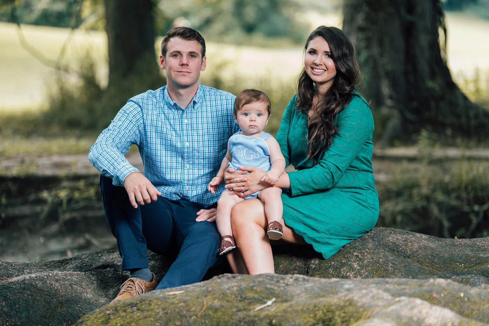 Family sitting outdoors on a tree trunk, with a man in a blue checkered shirt, a woman in a green dress, and a baby in a light blue outfit.