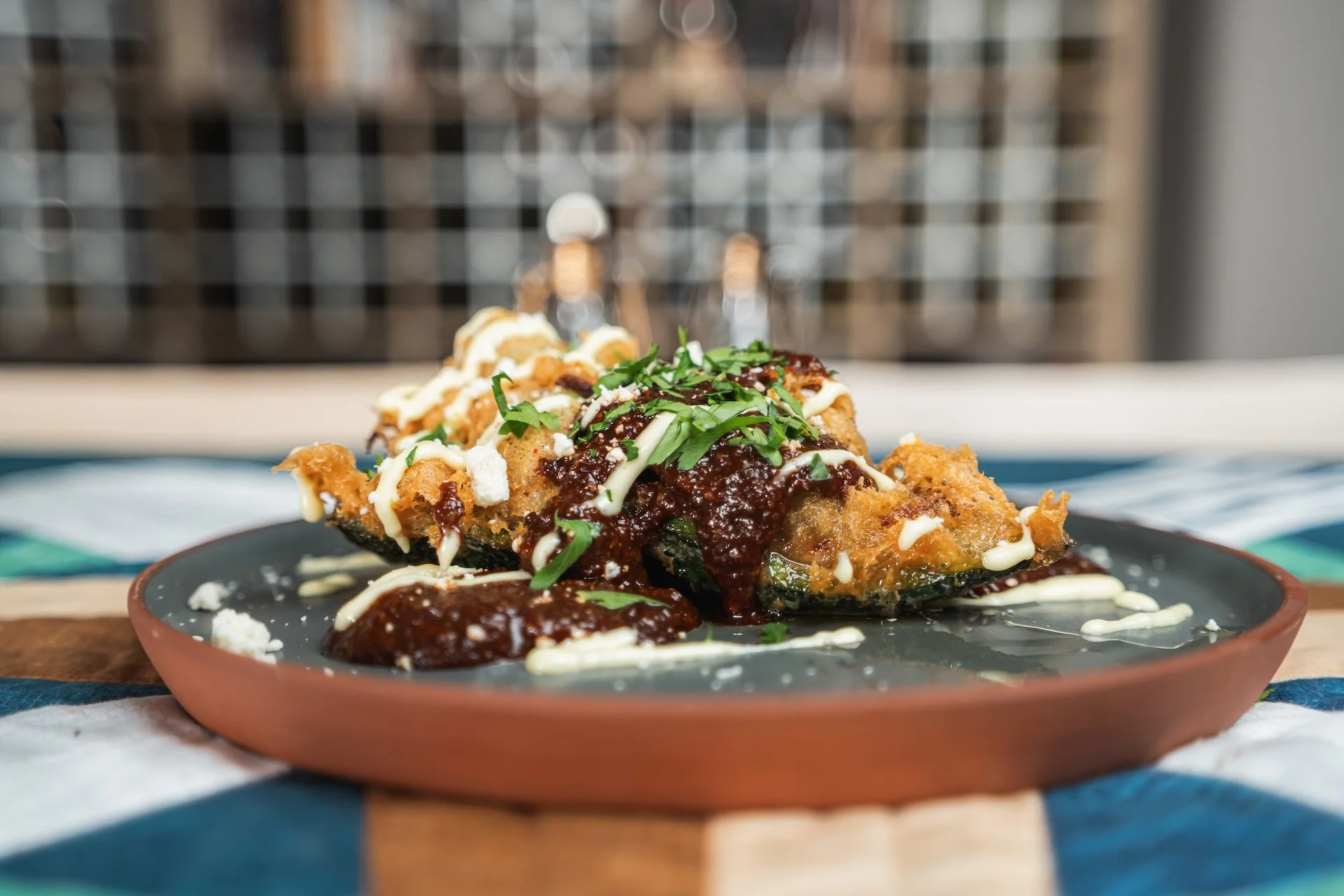 Fried fish fillet topped with dark sauce, drizzled with white sauce, garnished with chopped herbs, served on a dark plate.