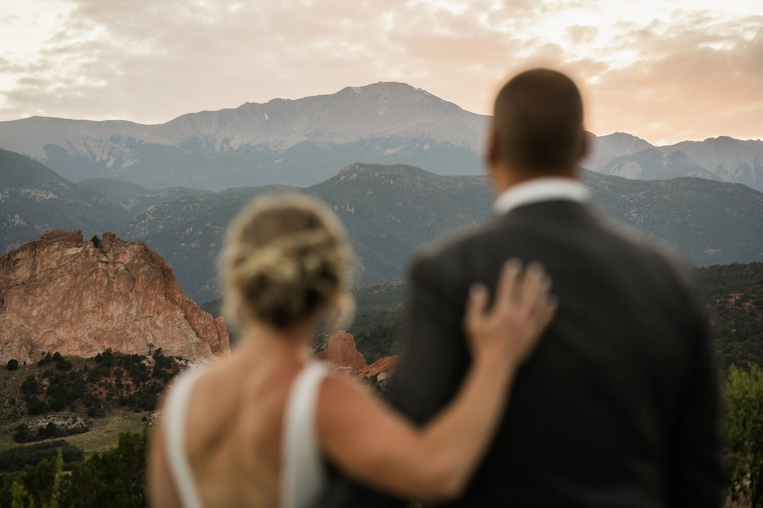 A couple stands with their backs to the camera, overlooking a mountainous landscape during sunset with rocky formations and lush greenery.