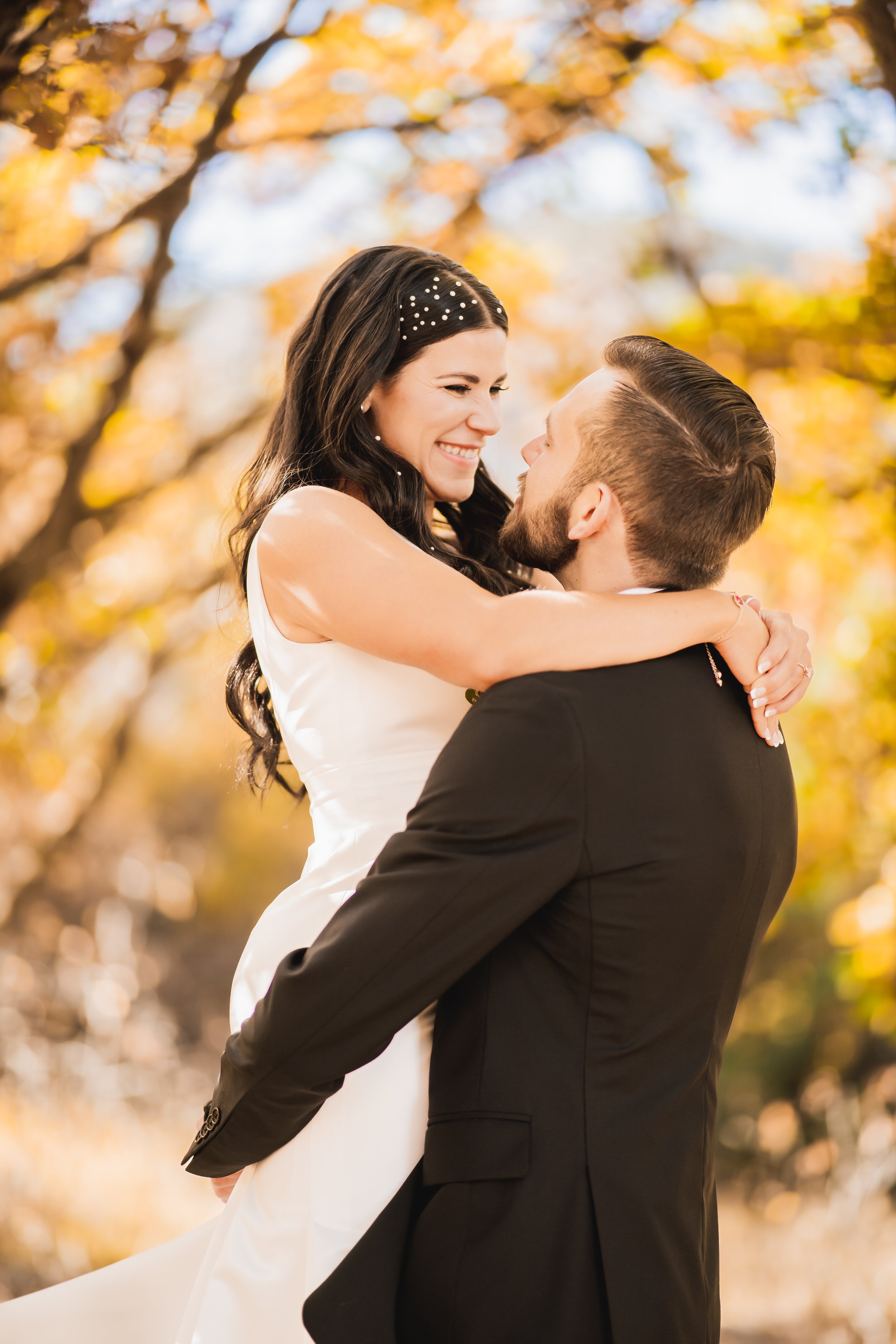 A couple embracing in a warm, sunlit outdoor setting with autumn foliage in the background. The woman wearing a white dress smiles at the man in a dark suit.