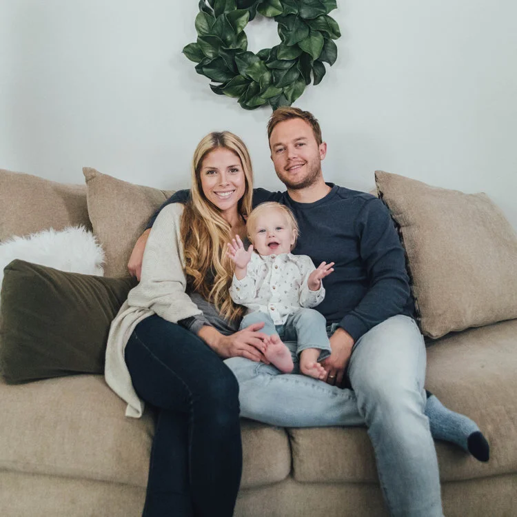 A family sitting on a beige couch, consisting of a man, a woman, and a baby. A green decorative wreath hangs on the wall behind them.