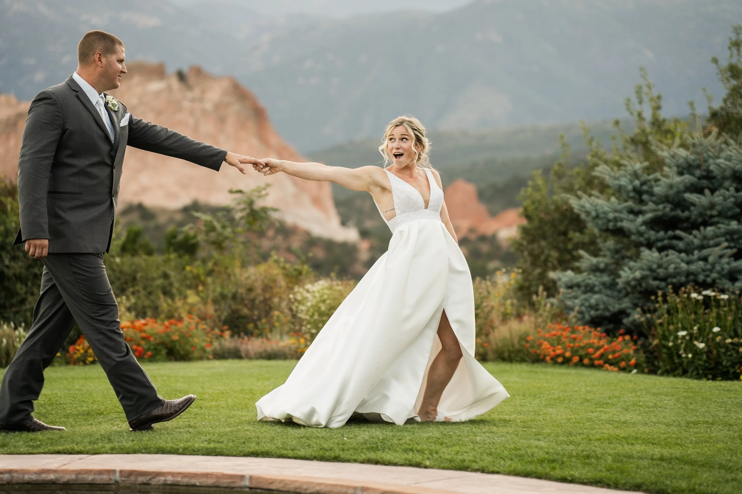 A bride and groom holding hands, walking on grass with rocky mountains in the background.