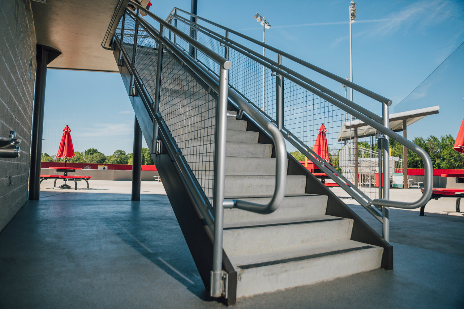 Modern outdoor stairs with metal railing at a recreational facility with red picnic tables and umbrellas in the background.