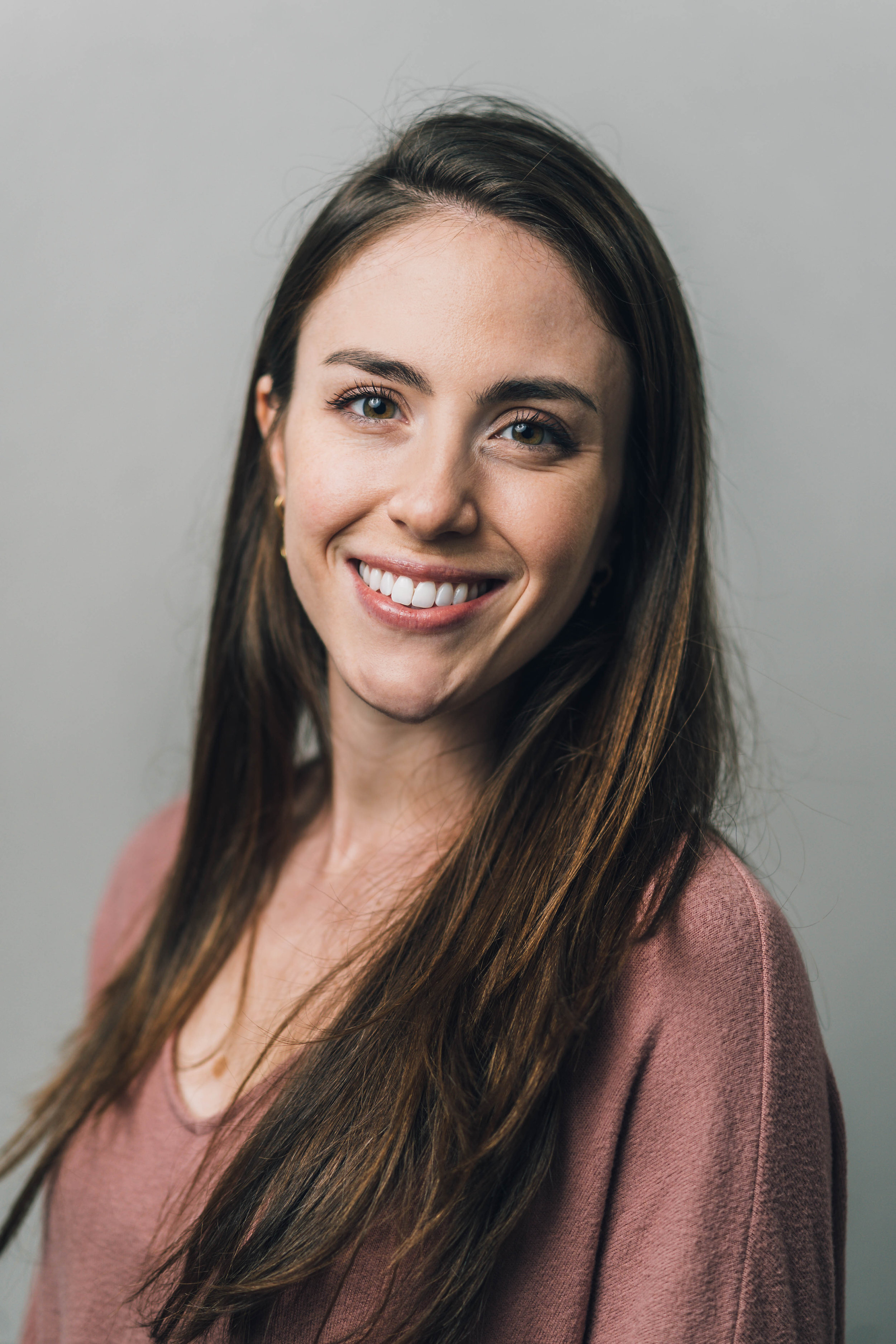 Smiling woman with long brown hair wearing a pink shirt against a plain background.
