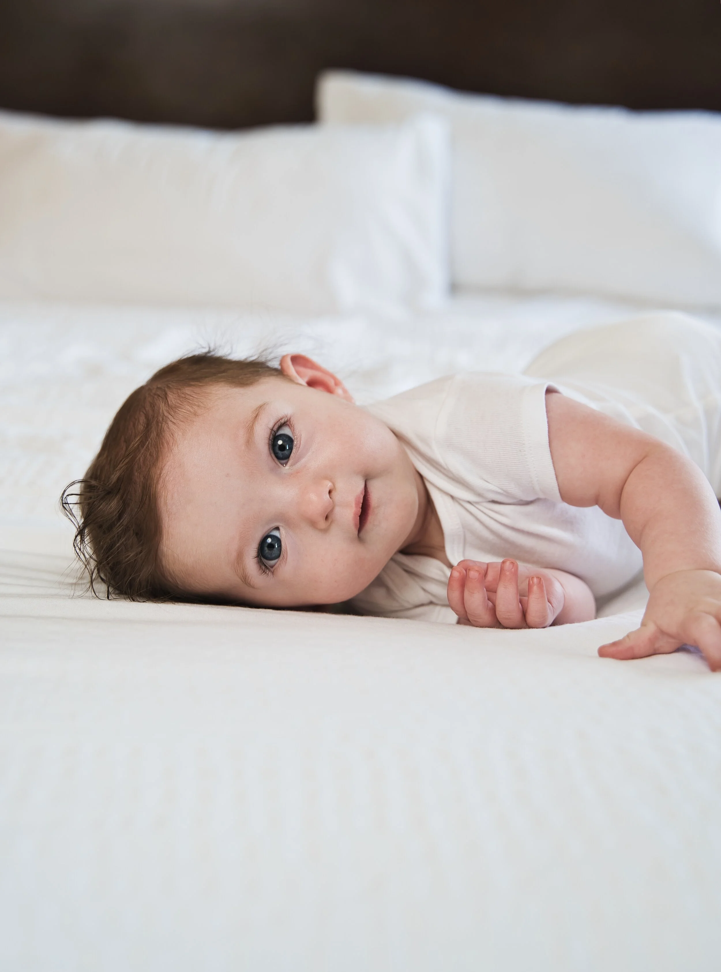 A baby in a white onesie lying on a bed with white sheets, looking towards the camera.