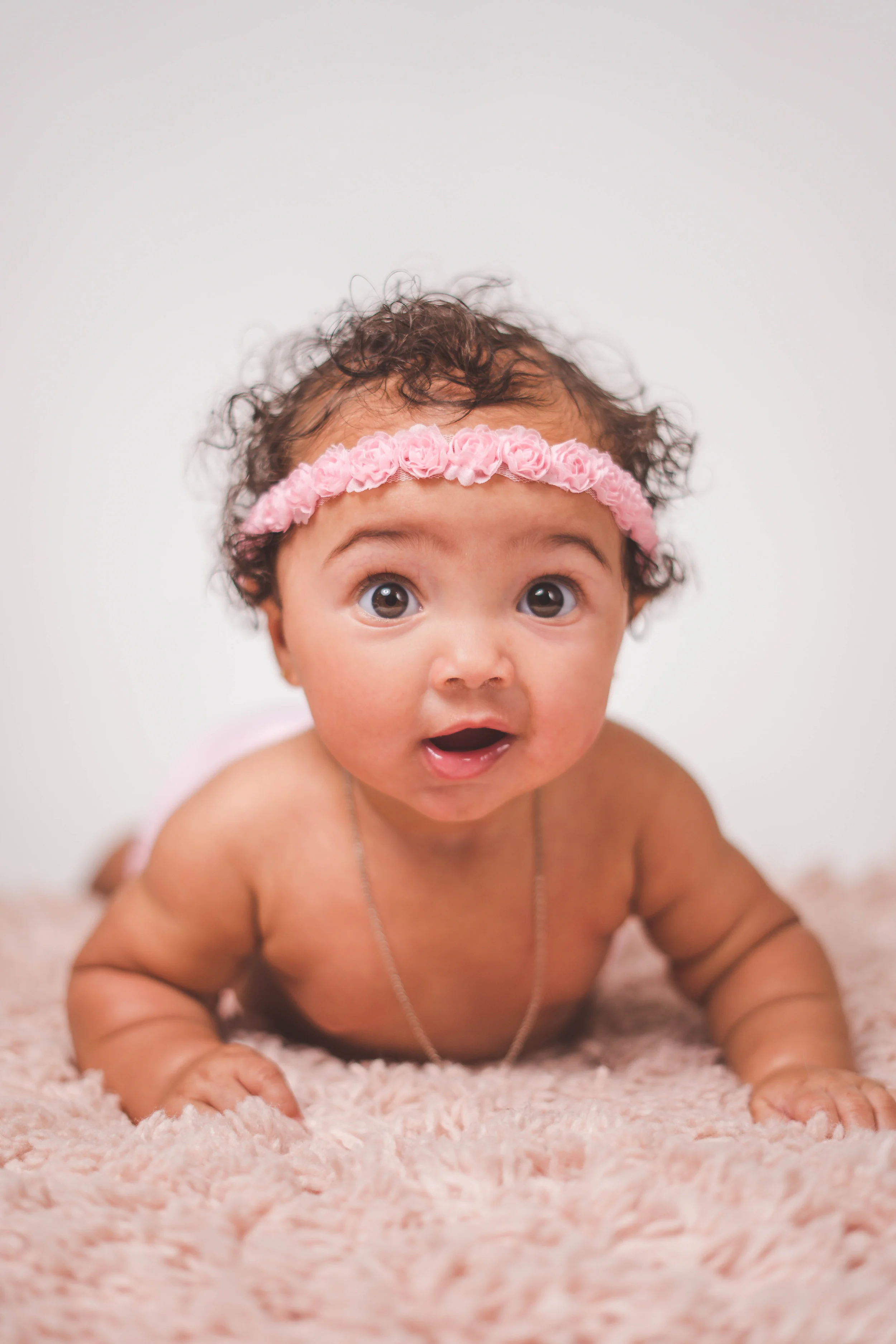 A baby wearing a pink floral headband lying on a soft pink carpet, looking up with a curious expression.