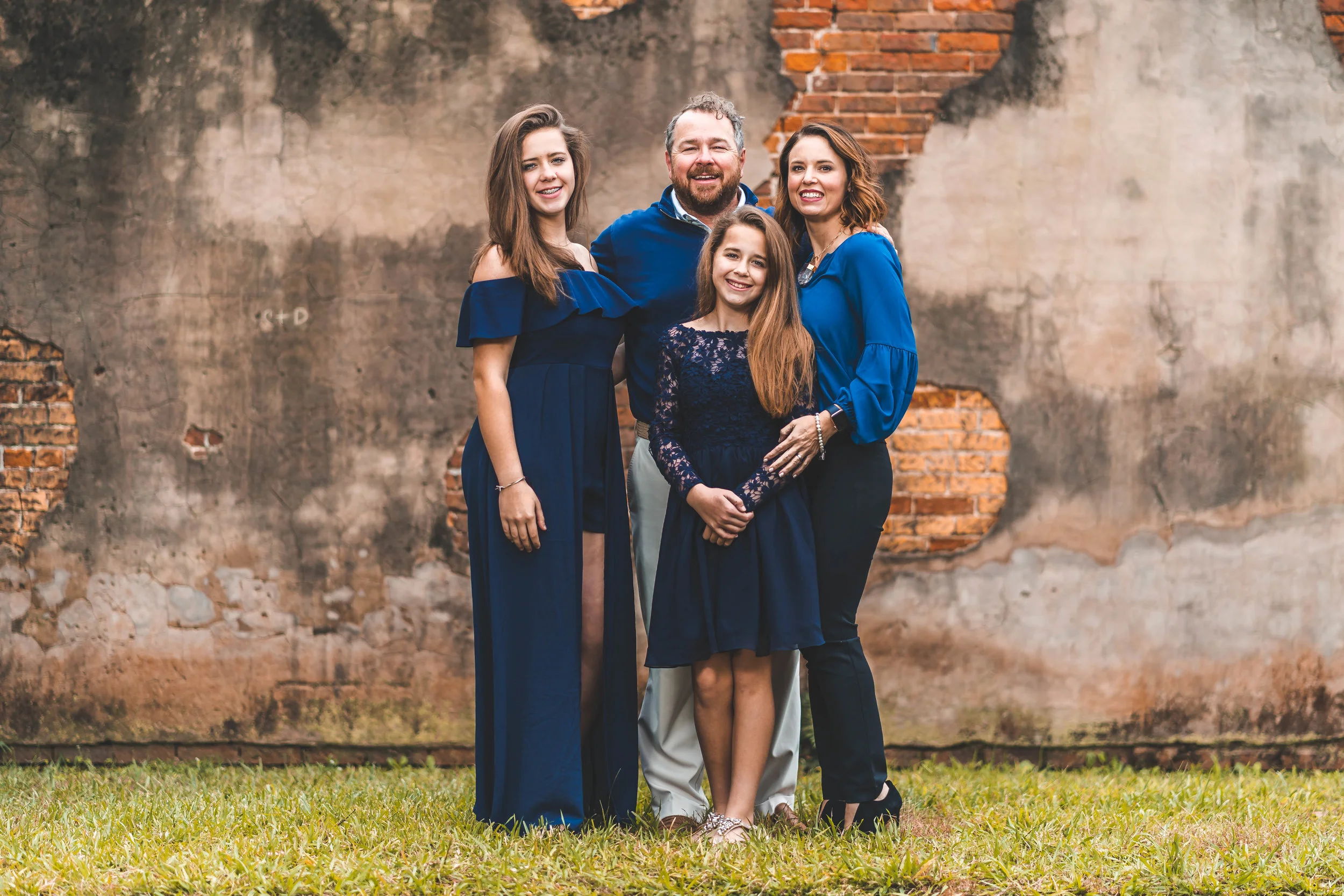 A family of four posing in front of an old brick wall; two adult women, one man, and a young girl wearing blue outfits.