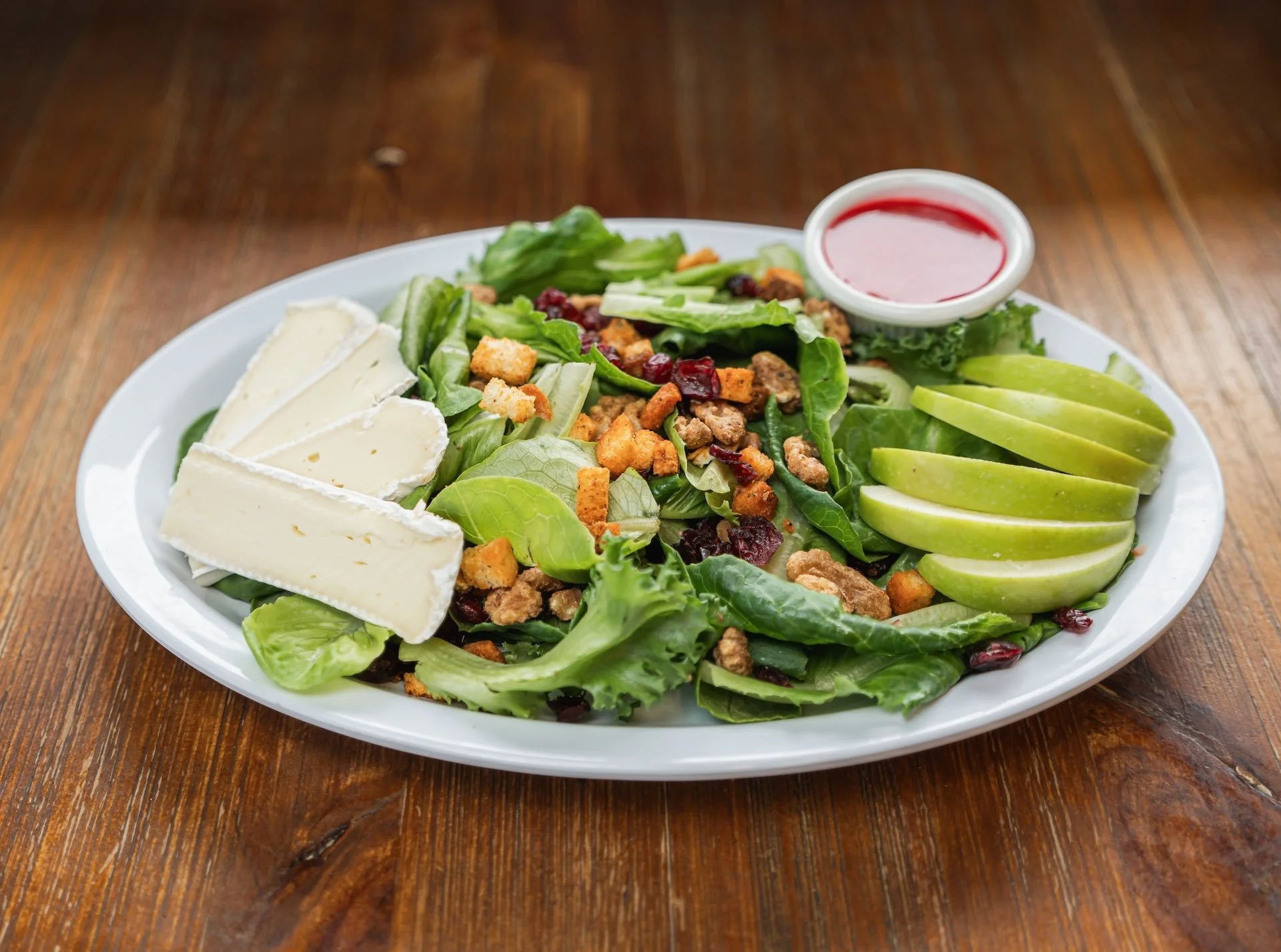 A white plate holding a salad with lettuce, slices of brie cheese, sliced green apple, crispy croutons, dried cranberries, ground meat, and a side of red vinaigrette dressing in a small dish, set on a wooden table.