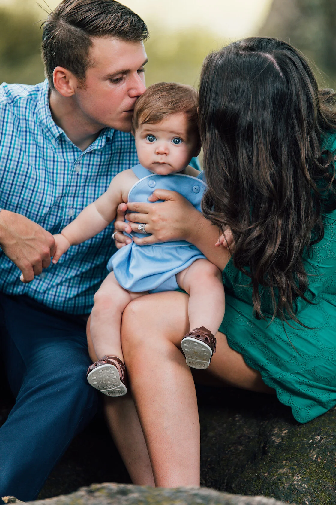 A family scene featuring a baby in a blue outfit sitting on a woman's lap, with a man kissing the baby on the head and holding its hand, outdoors.