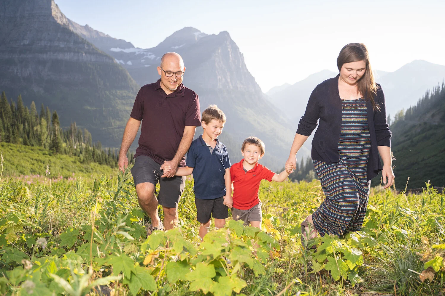 Family of four walking in a lush mountain landscape with towering peaks in the background.