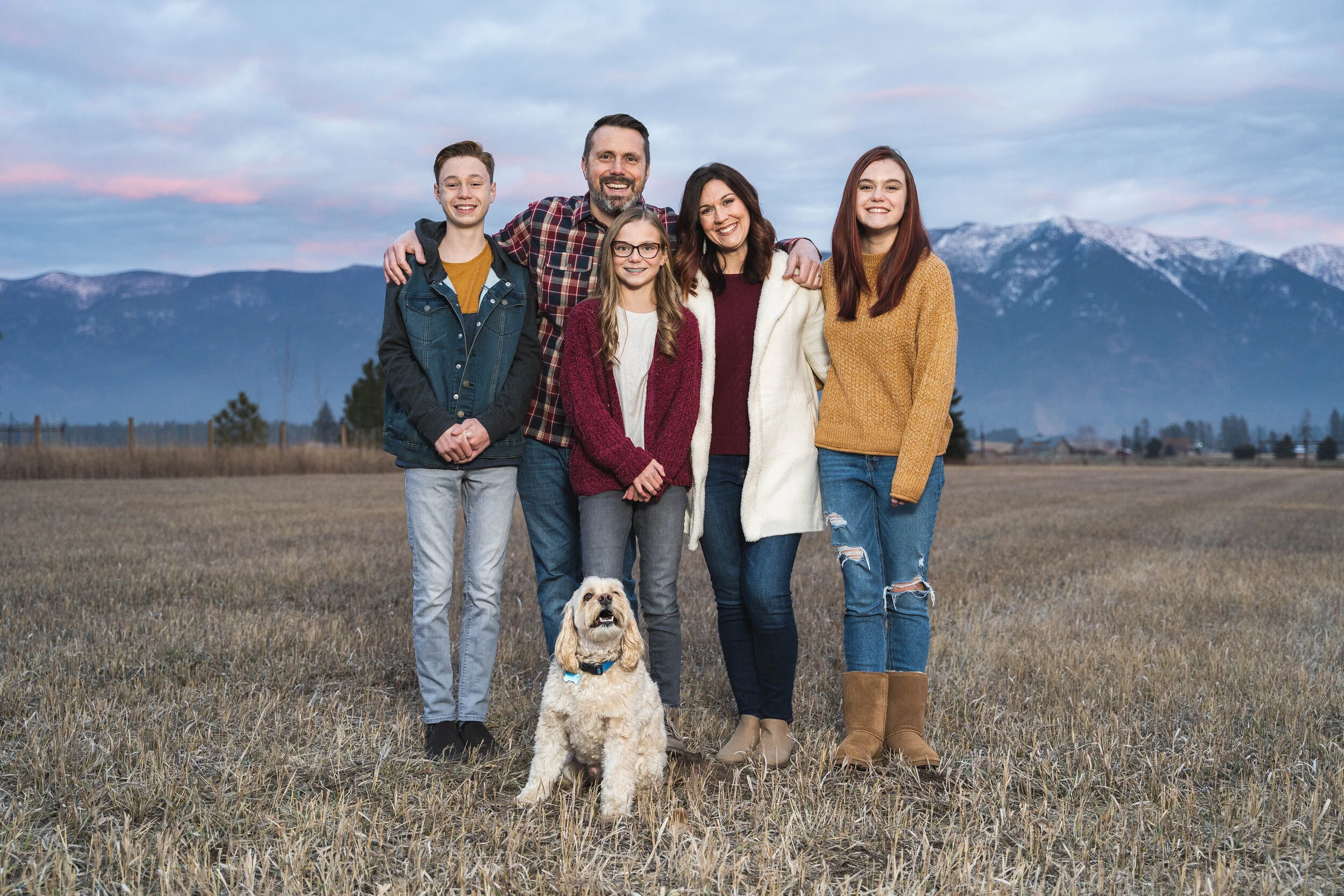 A family of five standing in an open field with a dog in front, mountainous landscape in the background.